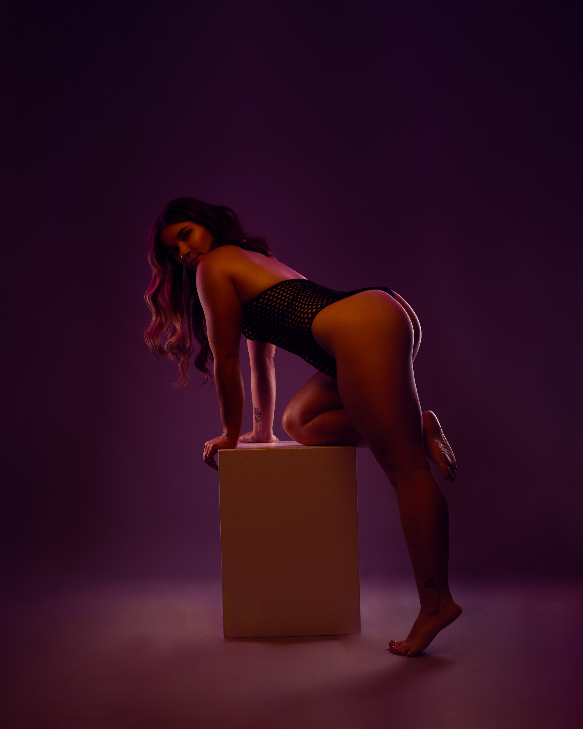 Fine art boudoir photography by Amanda Chantee featuring a woman leaning over a pedestal in black lingerie under purple and pink lighting.