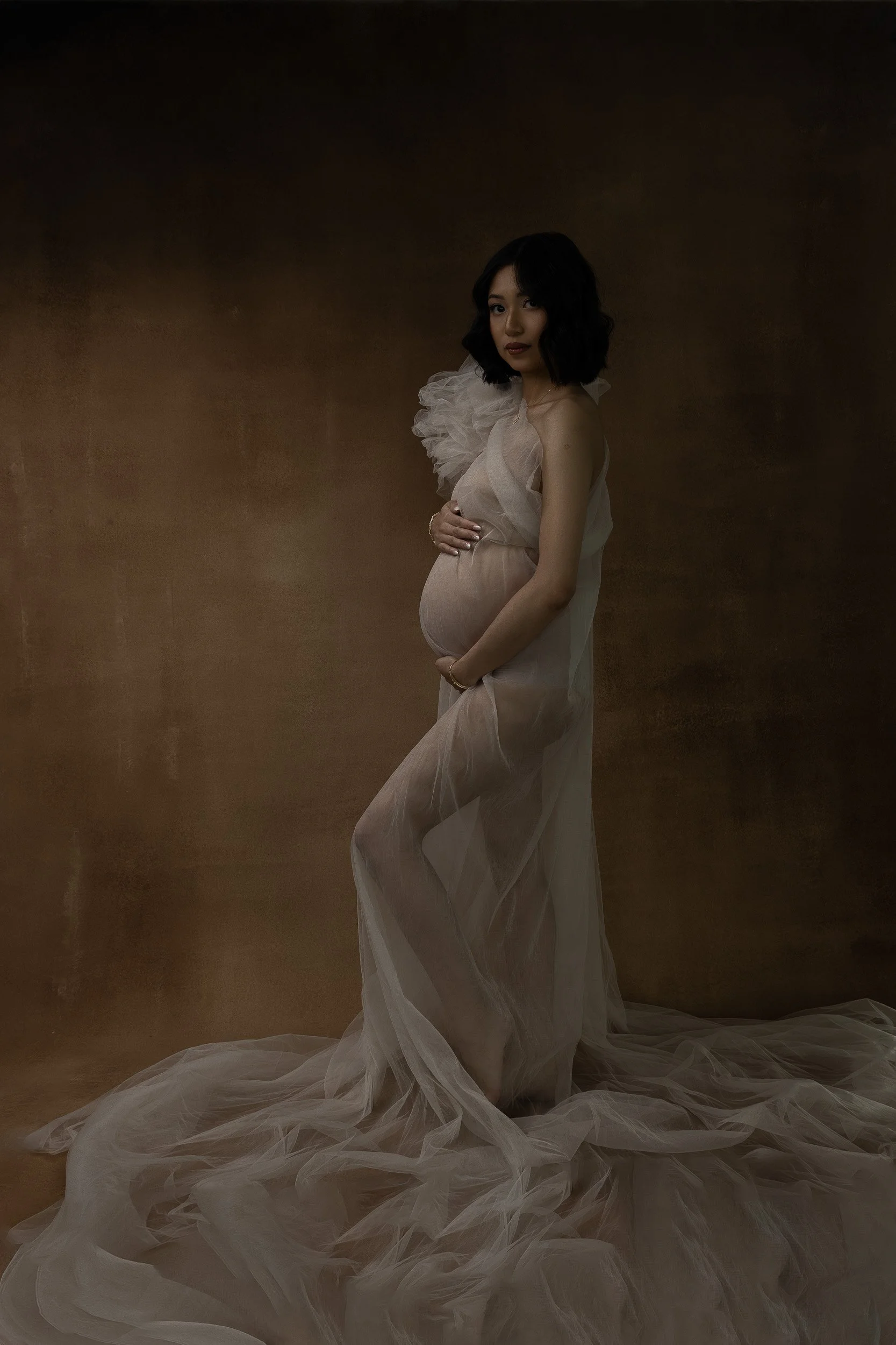 Fine art studio maternity portrait of an expectant mother in a sheer ivory gown, cradling her belly against a warm brown backdrop.