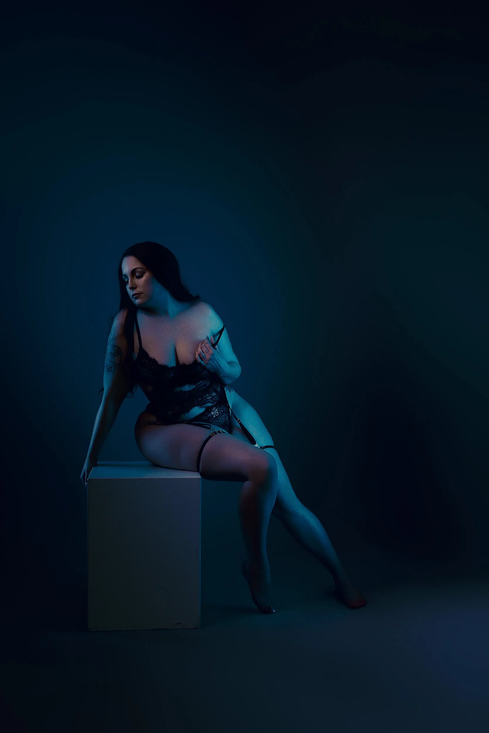Vancouver WA boudoir portrait of a woman seated in black lingerie on a pedestal beneath dramatic blue studio lighting.