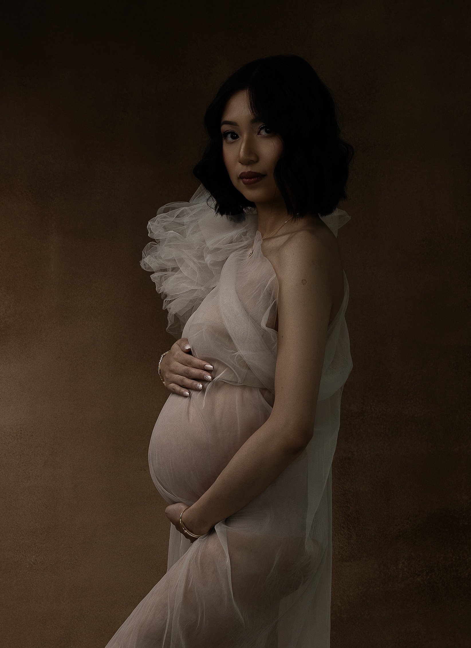 Fine art maternity portrait near Portland, Oregon, featuring a pregnant woman in a soft neutral gown against a warm painterly backdrop.