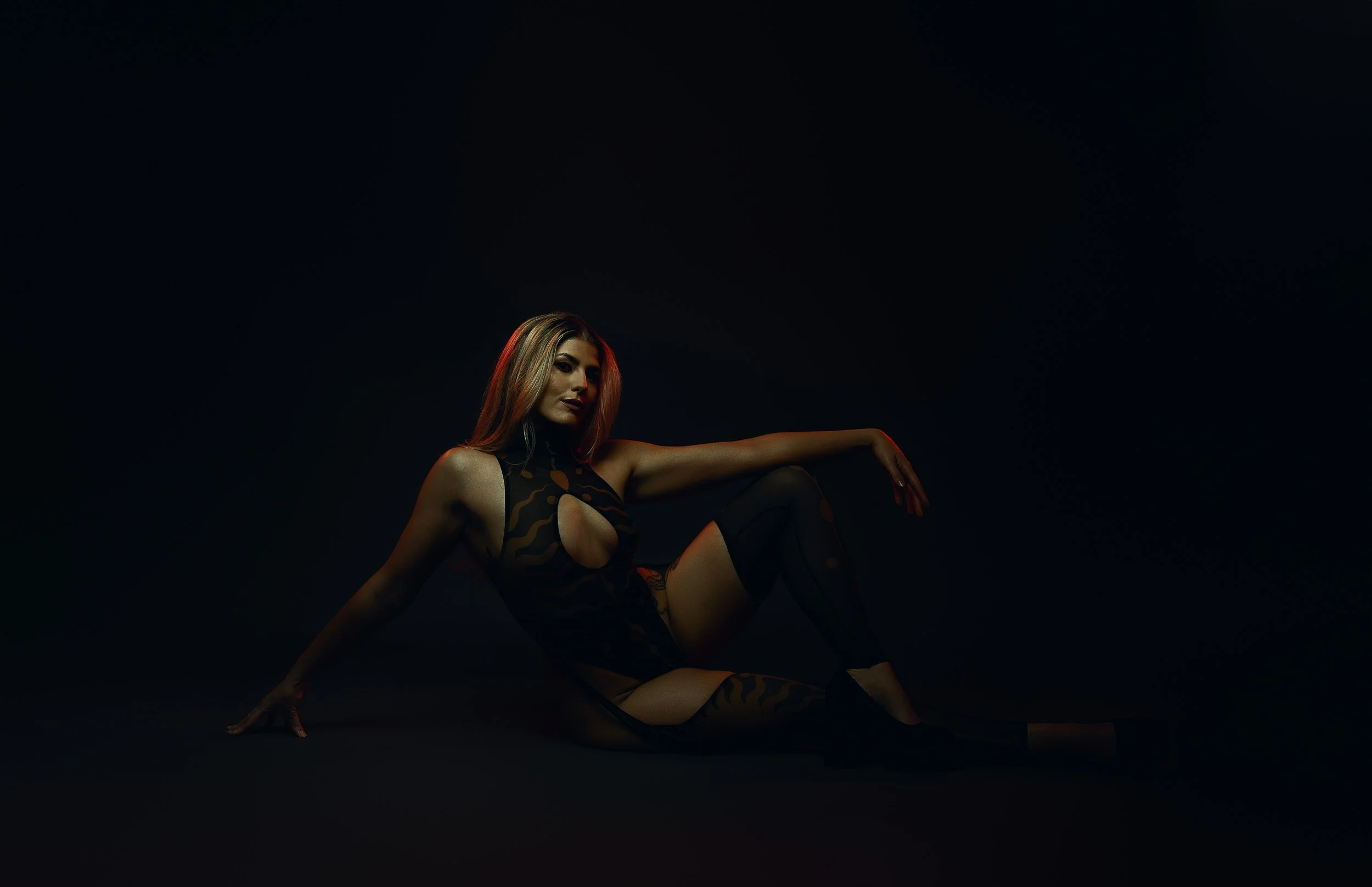 Woman seated on the floor in black lingerie under low-key dramatic studio lighting during a fine art boudoir session.