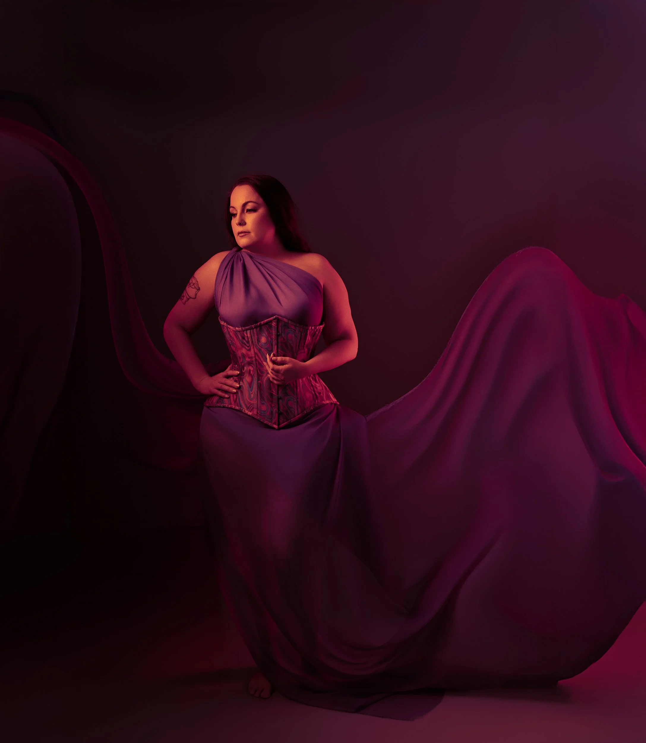 Luxury boudoir portrait of a woman in a corset framed by sweeping fabric under rich magenta lighting near Portland, Oregon.