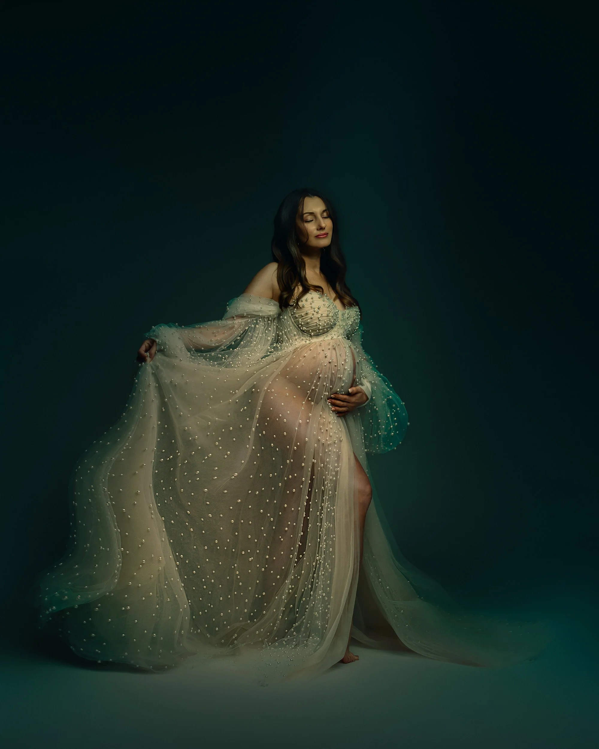 Fine art maternity portrait by Amanda Chantee featuring a pregnant woman in a sparkling sheer gown under moody teal studio lighting.