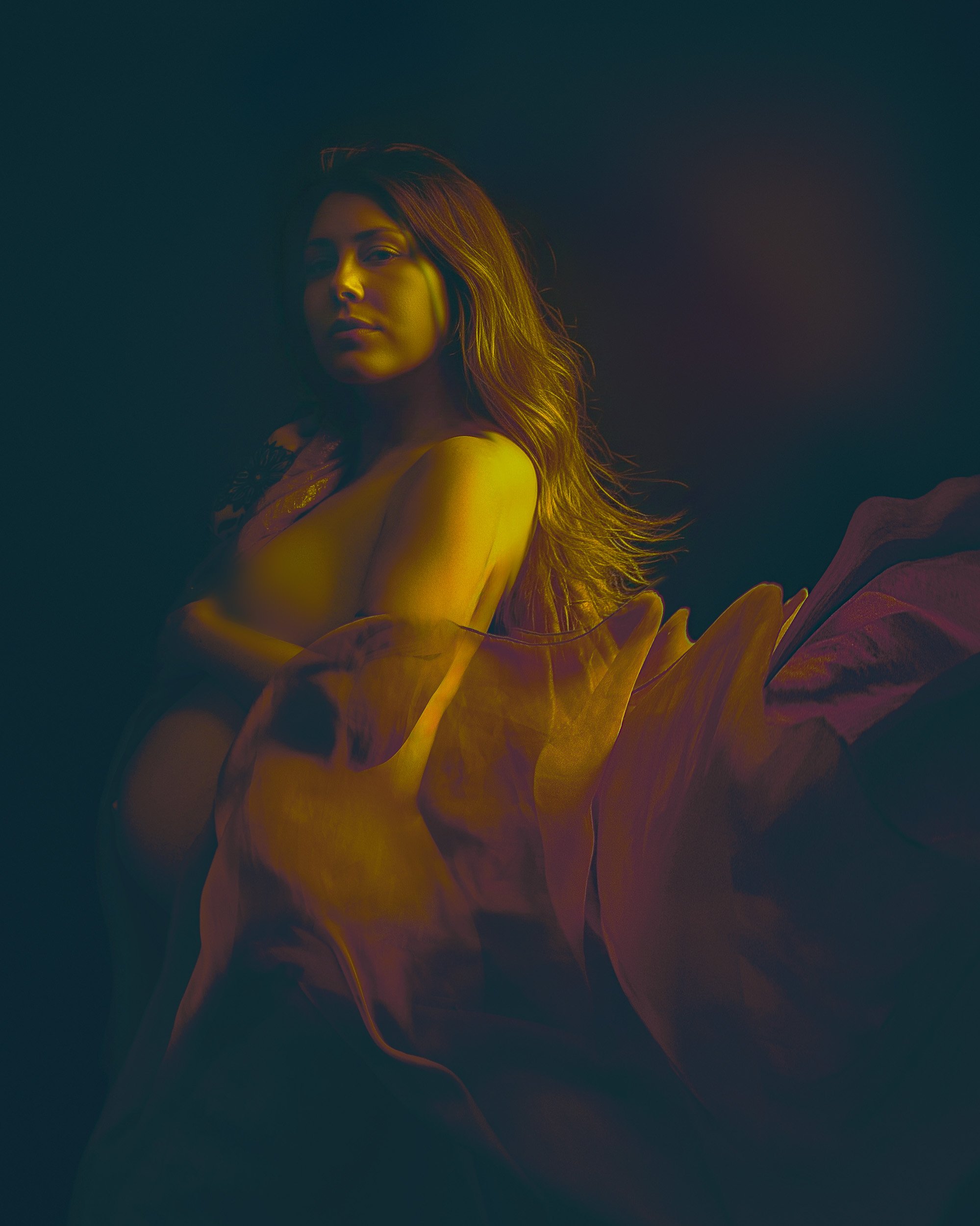 Artistic maternity portrait by Amanda Chantee near Portland, Oregon, featuring a pregnant woman wrapped in flowing fabric under rich amber and teal studio lighting.
