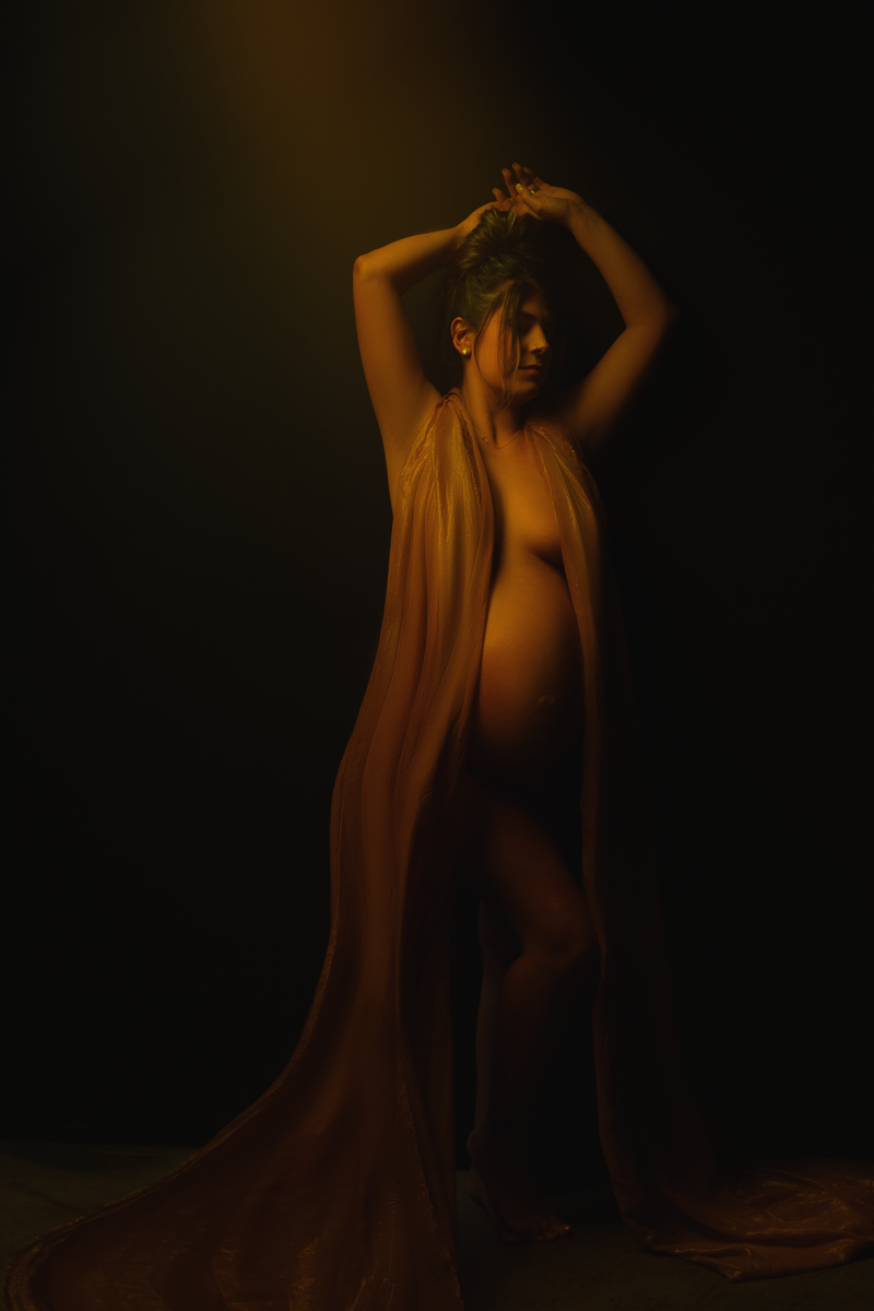 Fine art maternity portrait of a woman draped in sheer golden-brown fabric, posing against a dark background with warm studio lighting by Amanda Chantee