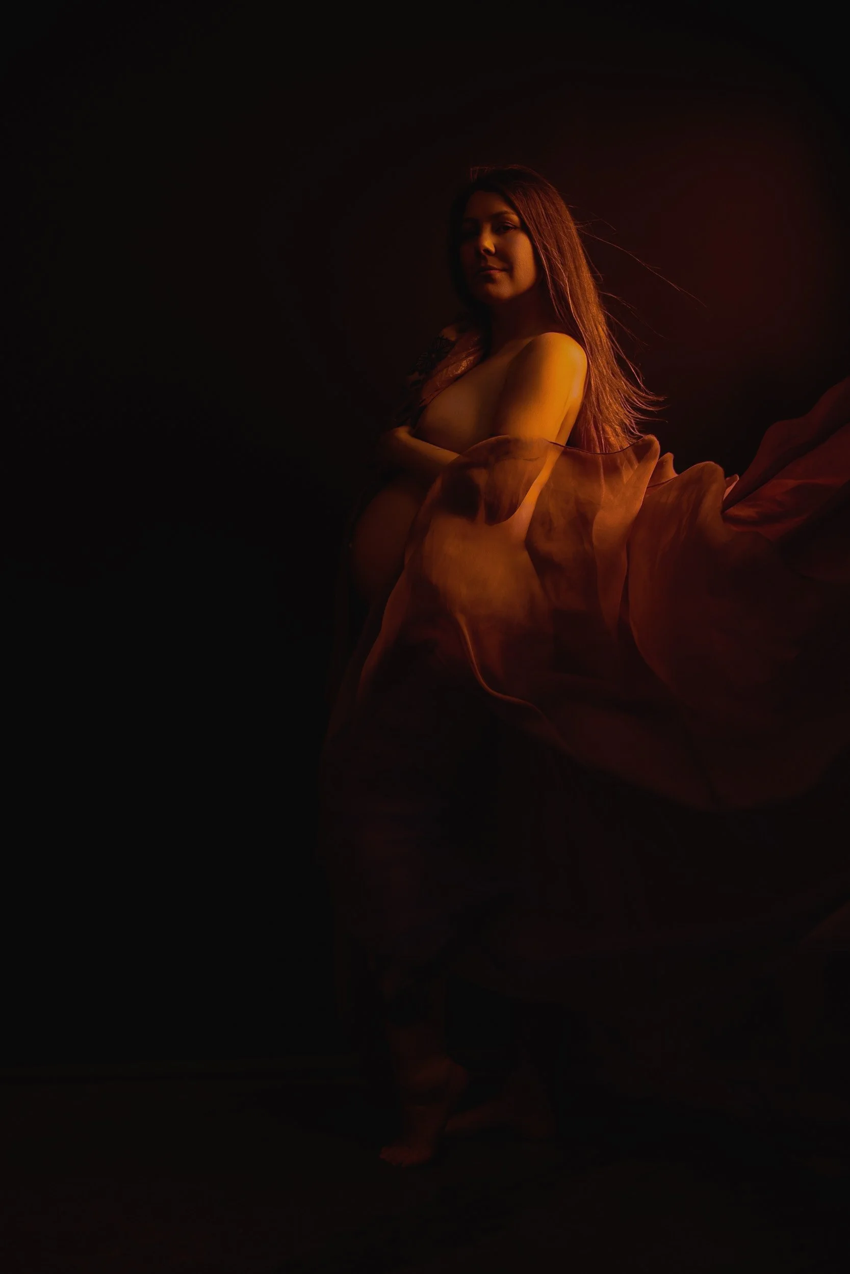 Moody fine art maternity photograph of a pregnant woman surrounded by flowing fabric, illuminated by warm amber studio lighting.