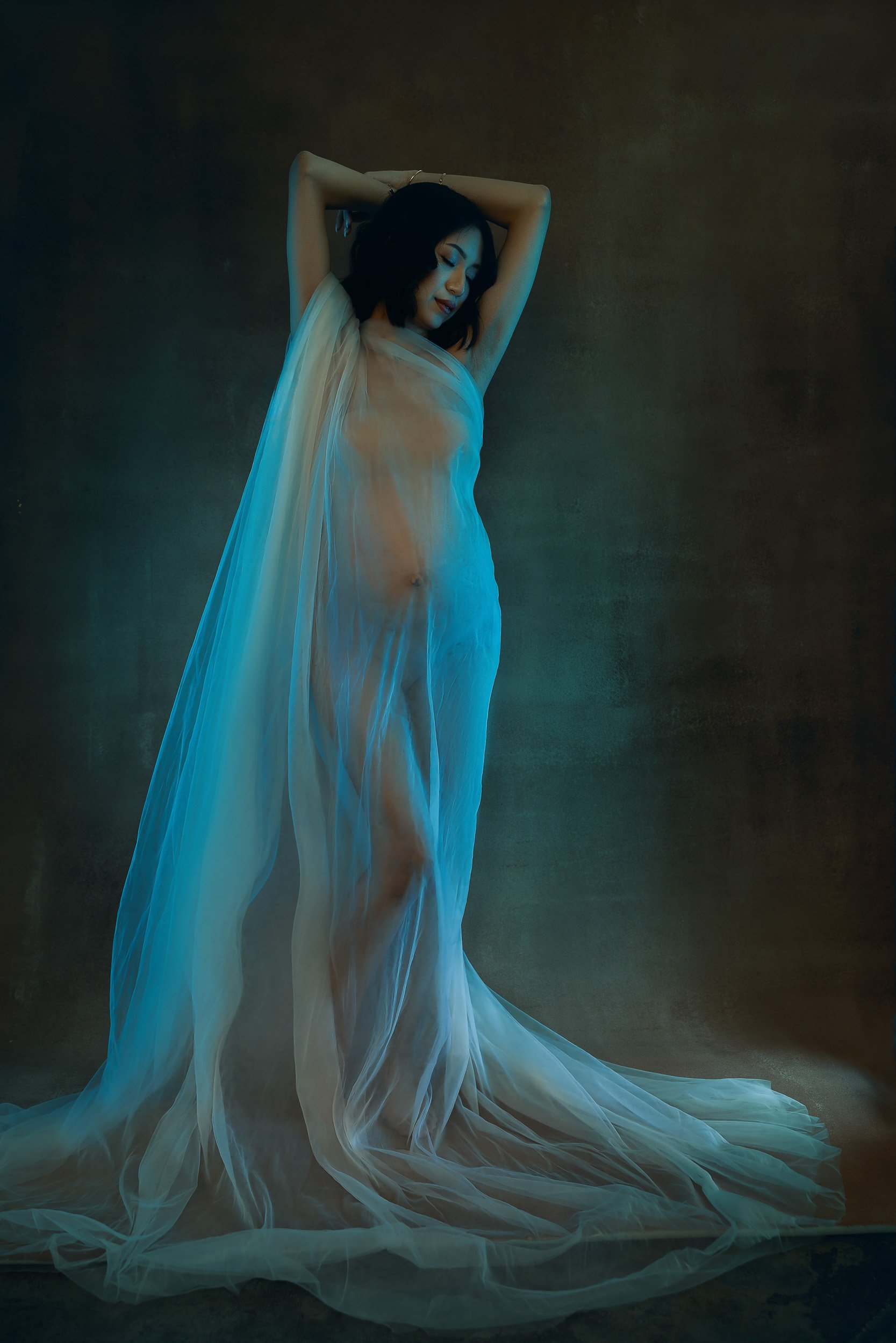 Artistic maternity portrait of a pregnant woman wrapped in sheer white fabric with soft teal lighting and an ethereal fine art look.
