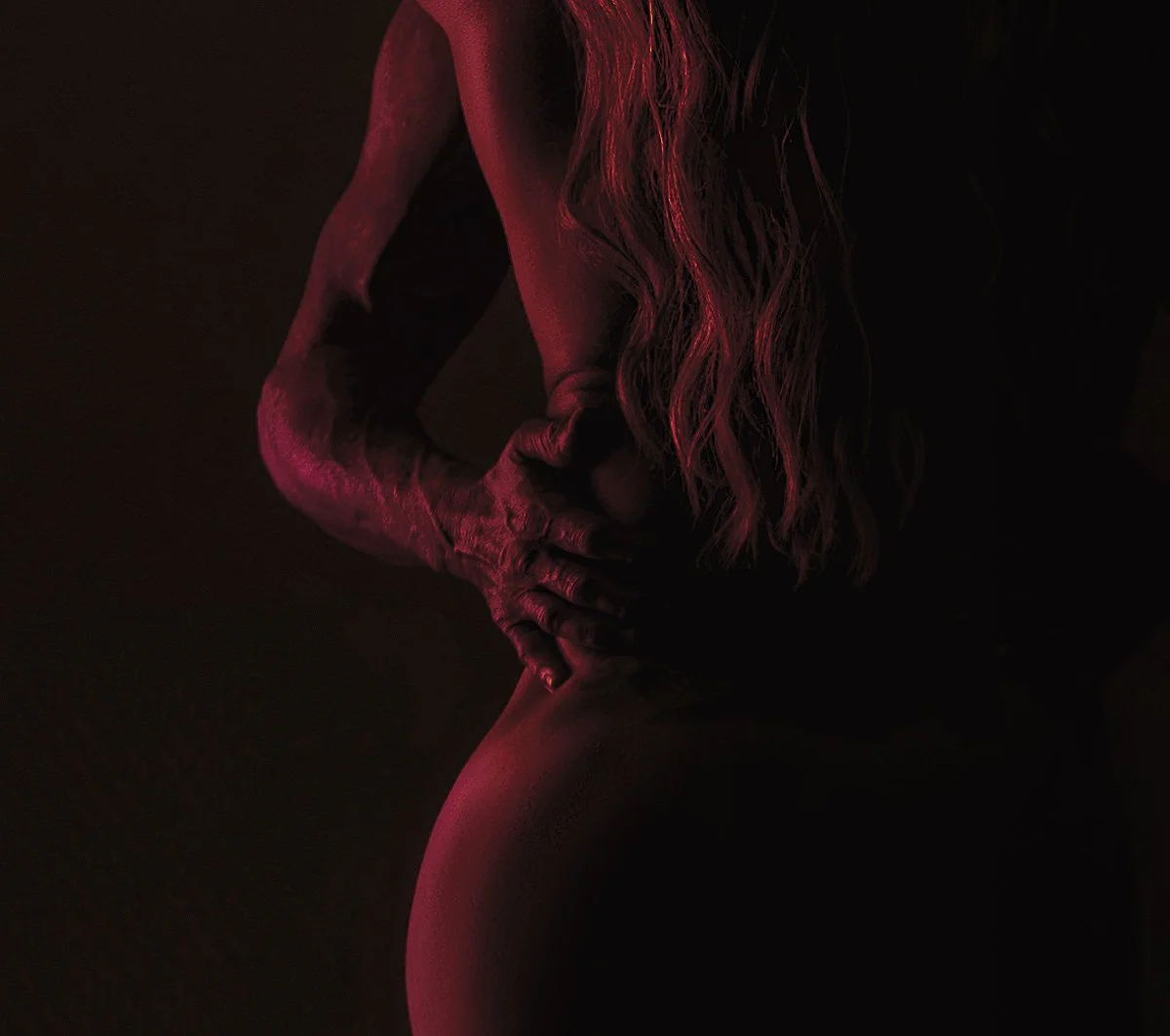 Close-up fine art boudoir portrait highlighting curves, shadow and body shape under dramatic red studio lighting.