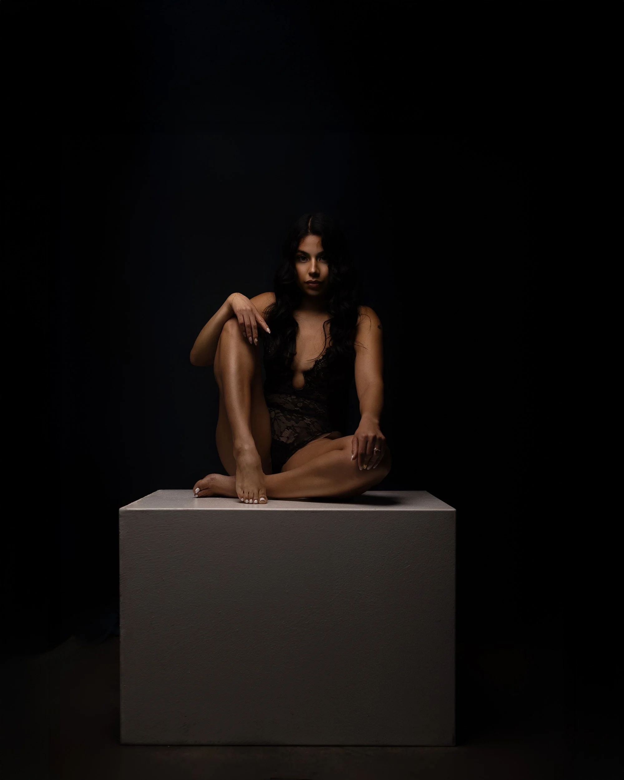Woman seated on a white pedestal in a dark studio portrait with sculptural posing and dramatic fine art lighting.