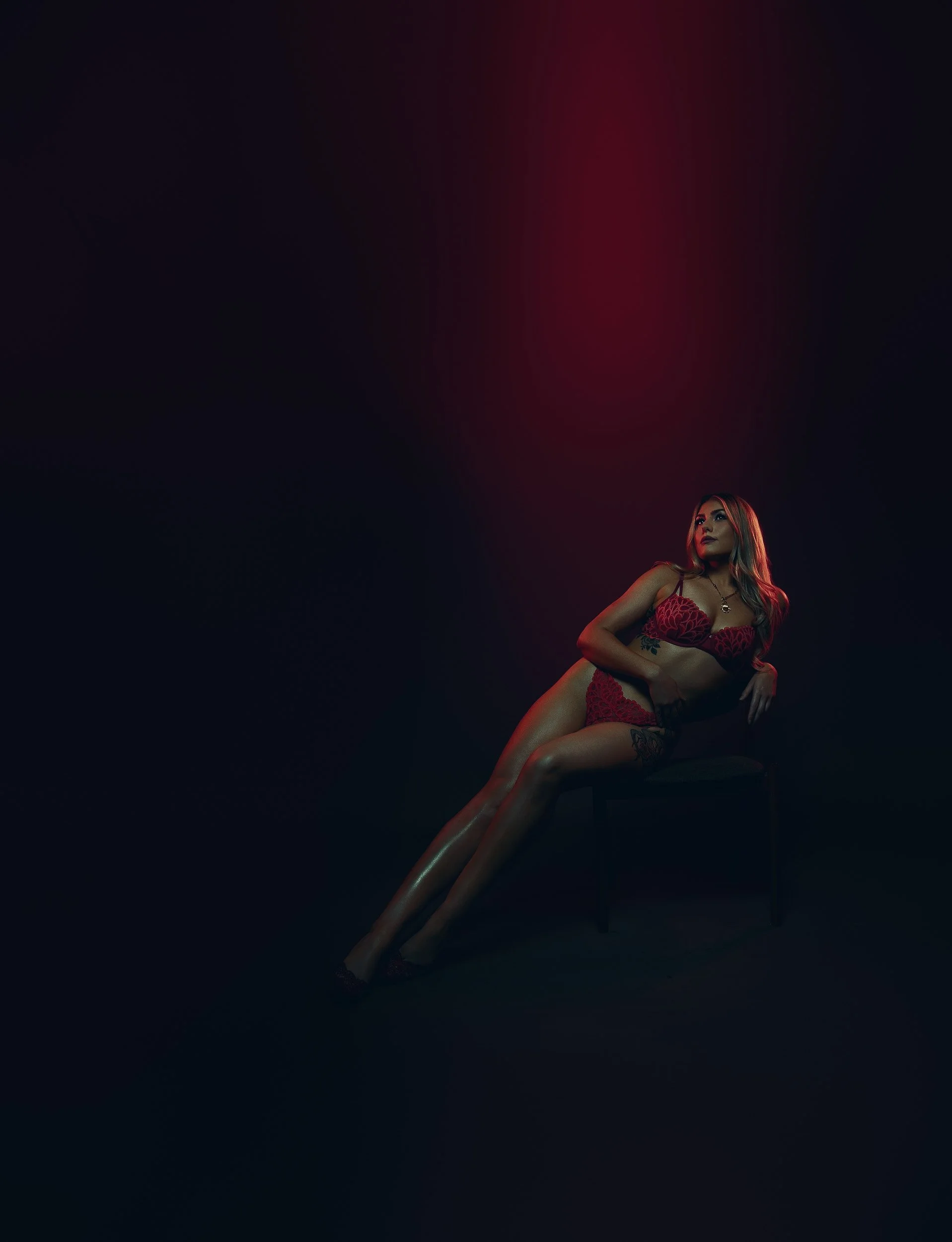 Woman reclining in red lingerie under moody red and blue lighting during a luxury boudoir photography session.