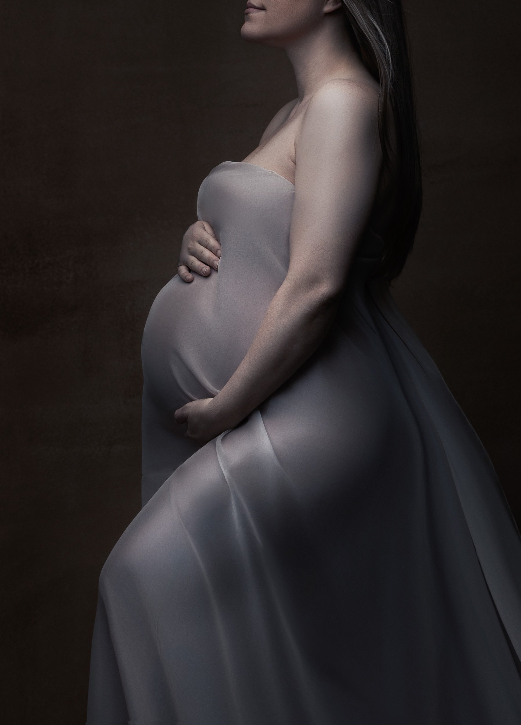 Close-up fine art maternity portrait highlighting a pregnant belly and fitted silver gown under soft sculptural lighting in Vancouver, WA.