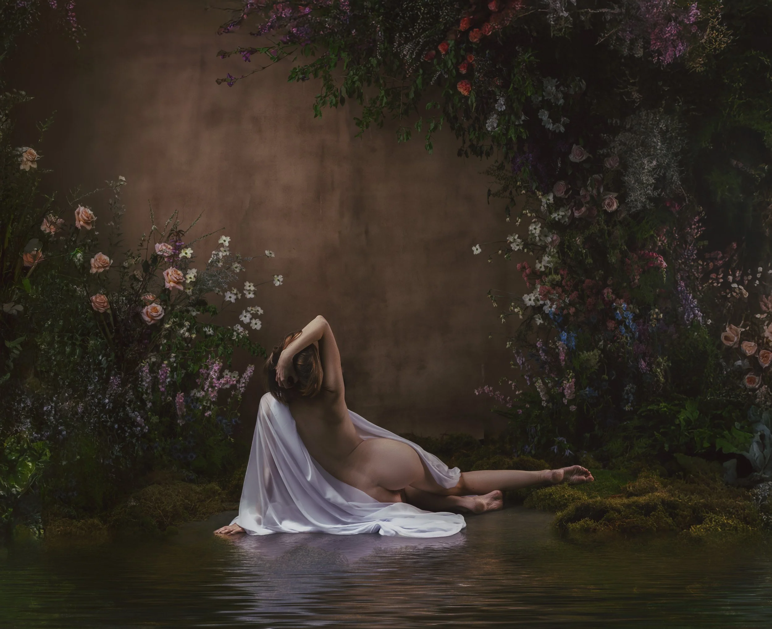 Fine art maternity portrait of a woman reclining in flowing white fabric within a romantic floral set.