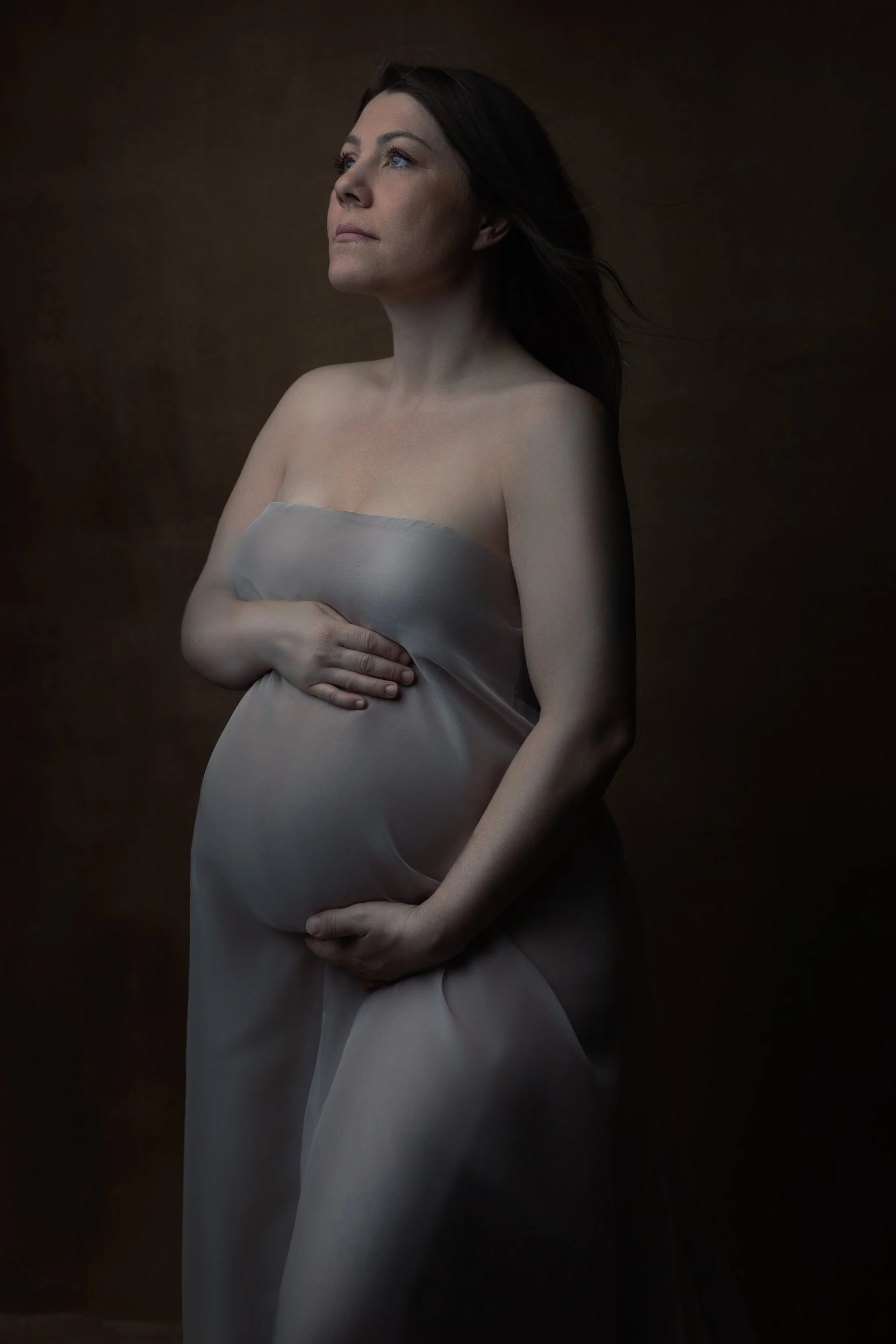 Close-up maternity portrait near Portland, Oregon, highlighting a pregnant belly in a fitted silver gown under soft sculptural studio lighting.