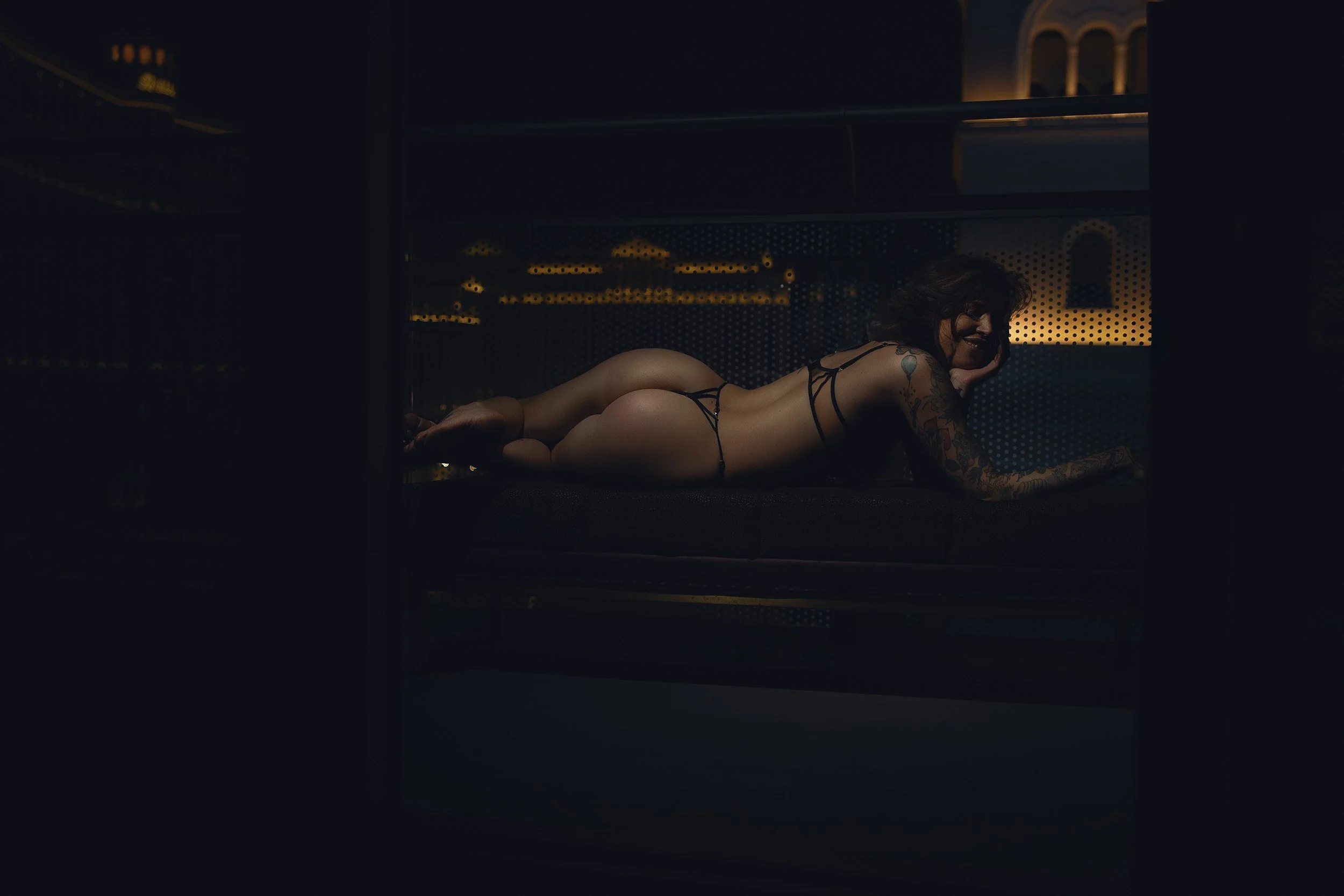 Tattooed woman lying on a dark sofa in strappy black lingerie during a moody fine art boudoir photography session in Vancouver, Washington.