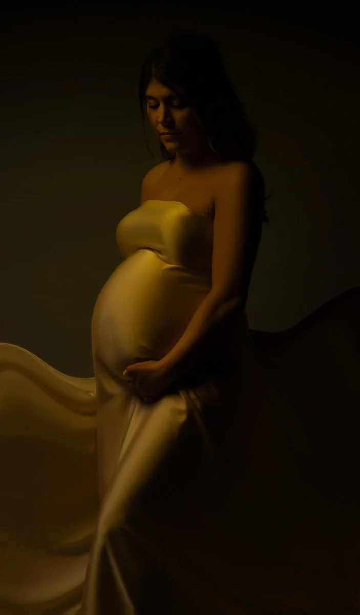 Luxury maternity photography near Portland, Oregon, featuring a pregnant woman in a flowing golden gown under soft dramatic studio light.