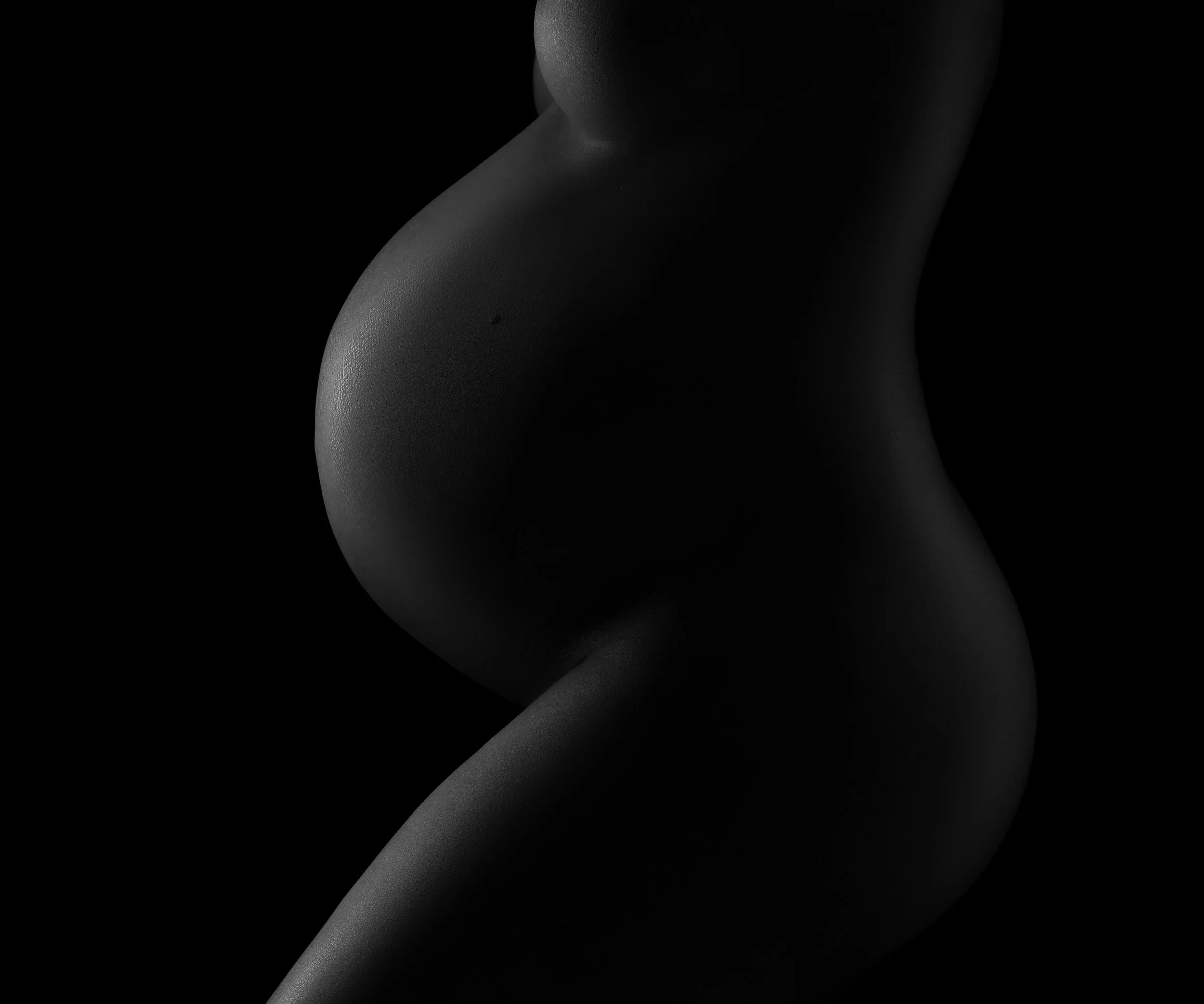 Close-up black and white maternity portrait highlighting the silhouette and curves of pregnancy with dramatic low-key lighting.