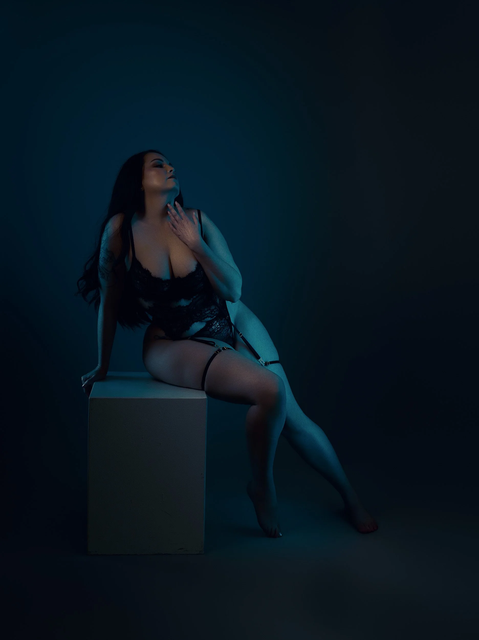 Editorial boudoir portrait by Amanda Chantee featuring a woman seated on a pedestal in black lingerie under moody teal light.