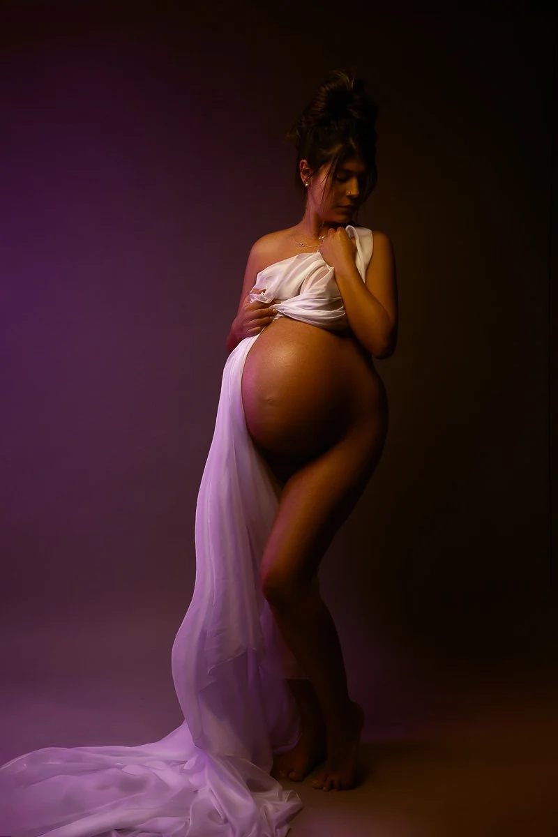 Elegant studio maternity portrait of an expectant mother draped in lavender fabric, posing against a rich purple backdrop.