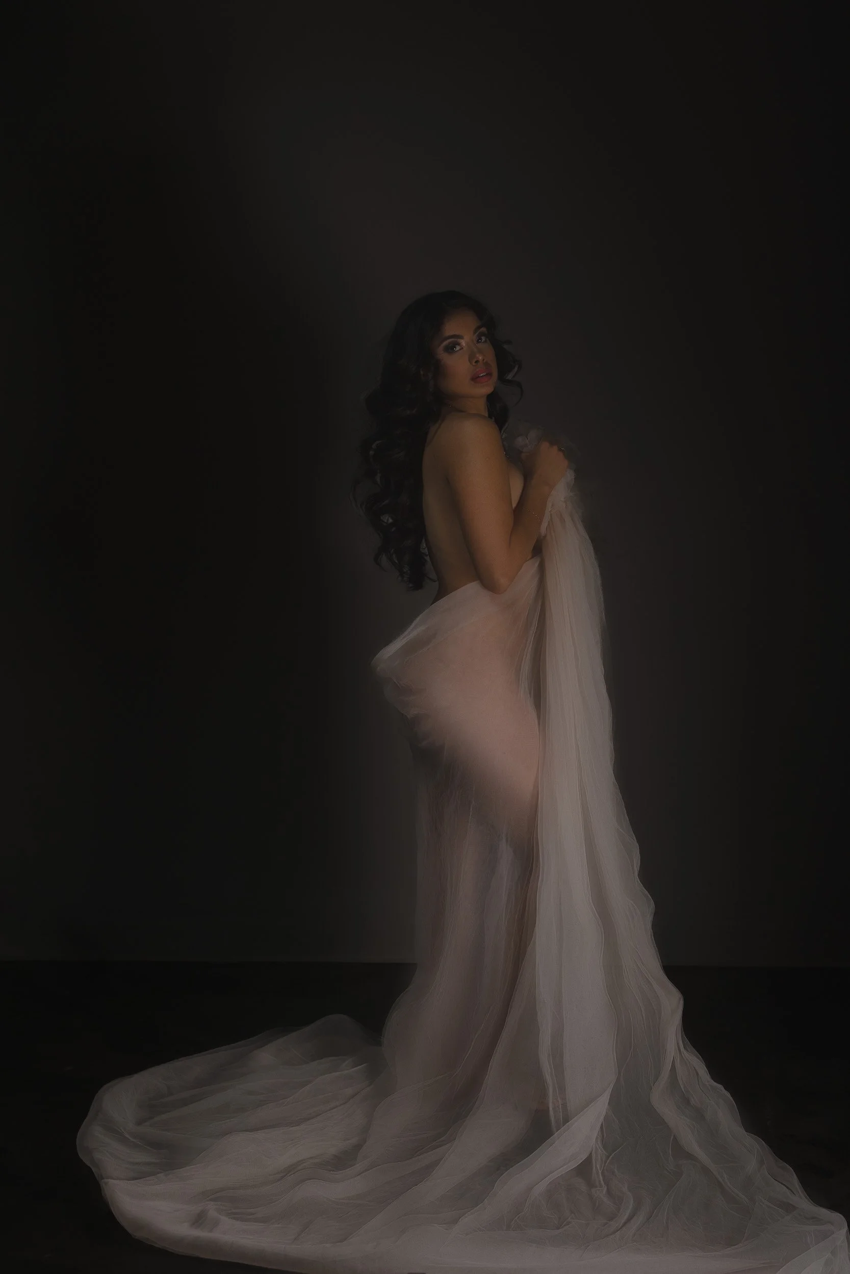 Artistic boudoir portrait by Amanda Chantee of a woman standing in flowing sheer white fabric with moody low-key lighting.