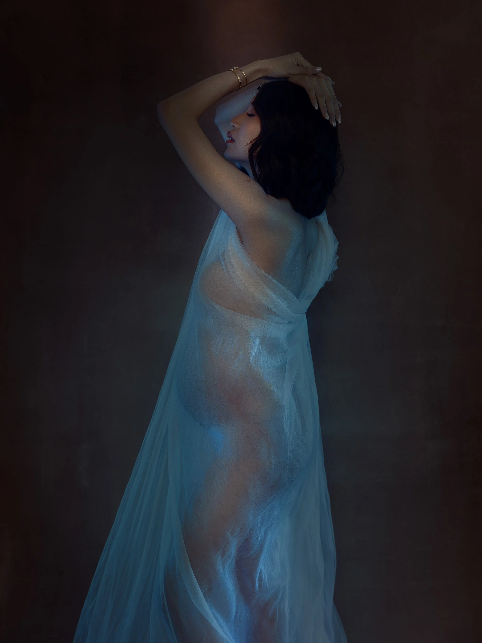 Artistic maternity portrait of a pregnant woman wrapped in sheer white fabric under soft blue studio lighting with an ethereal editorial feel.