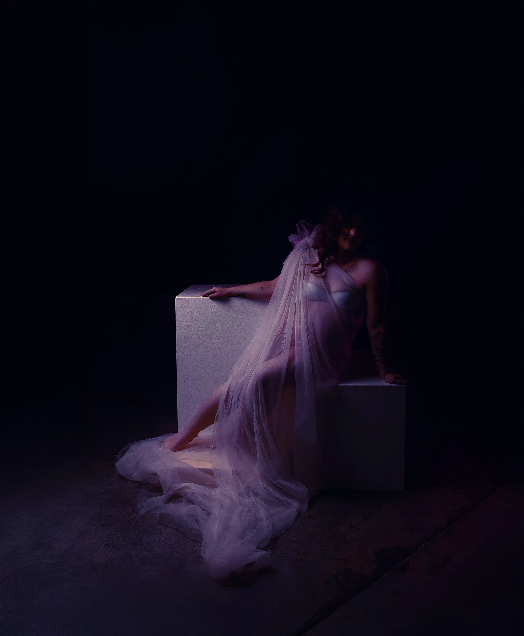 Moody maternity portrait near Portland, Oregon, featuring a pregnant woman reclining in a sheer lavender gown under soft purple studio light.