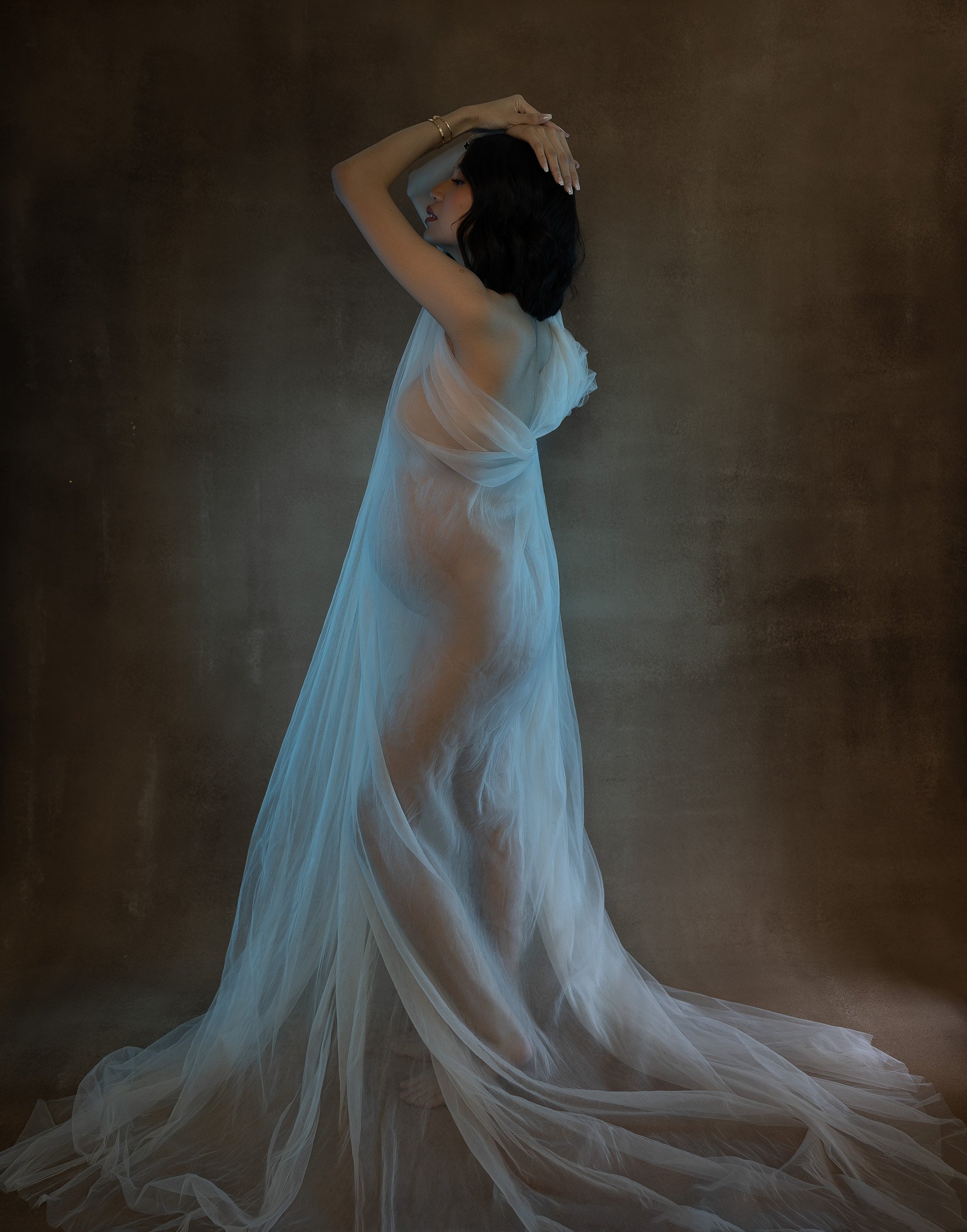 Full-length fine art maternity portrait of a pregnant woman posing in a flowing ivory gown against a textured brown studio backdrop.