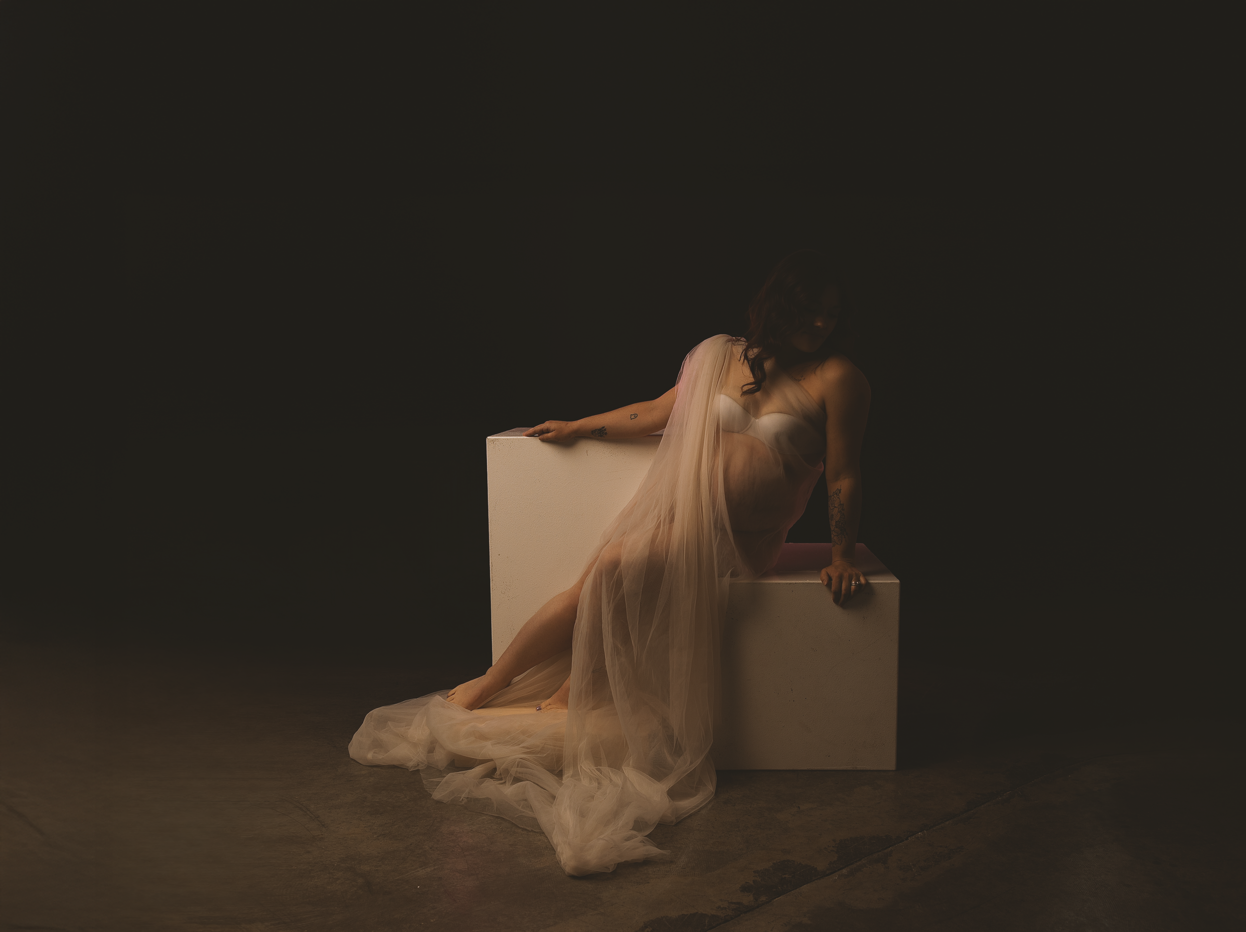 Fine art maternity portrait by Amanda Chantee in Vancouver, WA, featuring a pregnant woman draped in sheer white fabric and posed on a sculptural pedestal under warm low-key studio lighting.