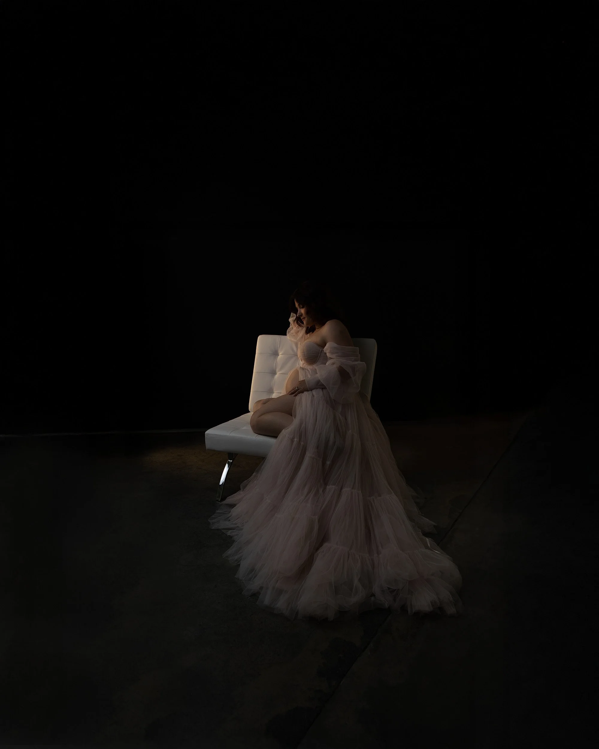 Editorial maternity portrait by Amanda Chantee featuring a pregnant woman seated in a voluminous gown against a dark studio background with cinematic lighting.