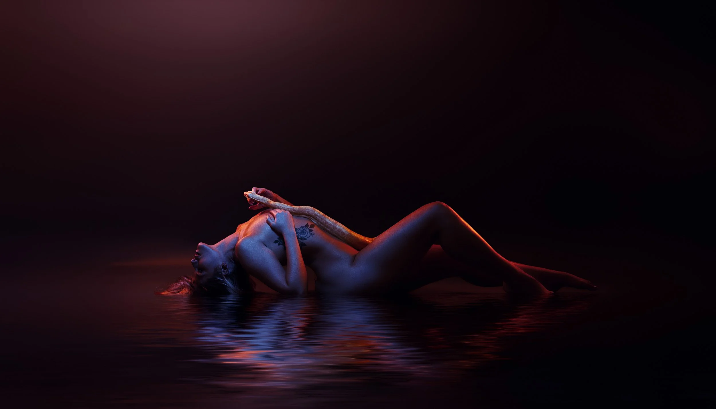 Woman reclining on a reflective floor in black lingerie with dramatic blue and red lighting during an artistic boudoir portrait.