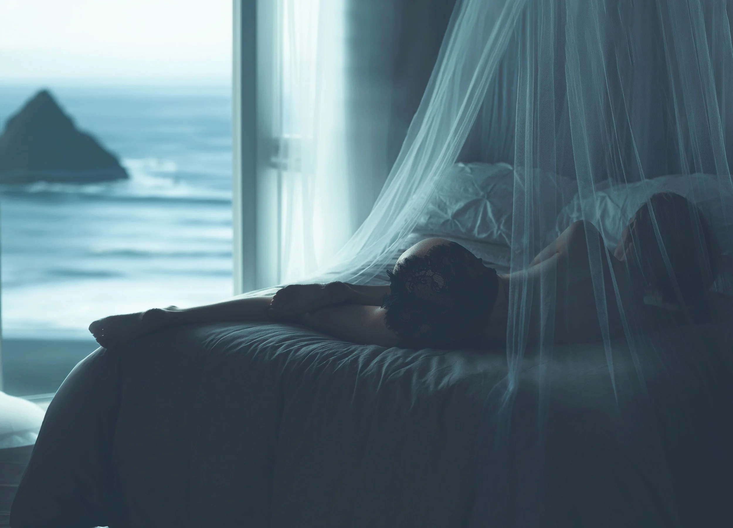 Intimate hotel boudoir portrait by Amanda Chantee of a woman reclining beneath sheer canopy fabric beside an ocean-view window.