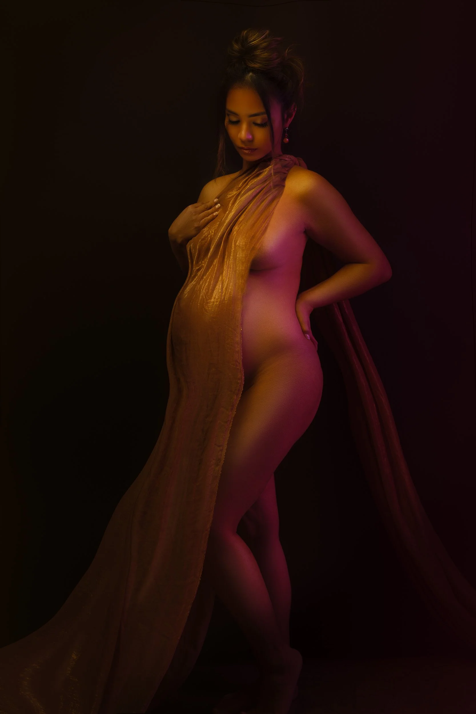 Fine art maternity portrait by Amanda Chantee in Vancouver, WA, featuring a pregnant woman draped in flowing gold fabric under dramatic warm studio lighting.