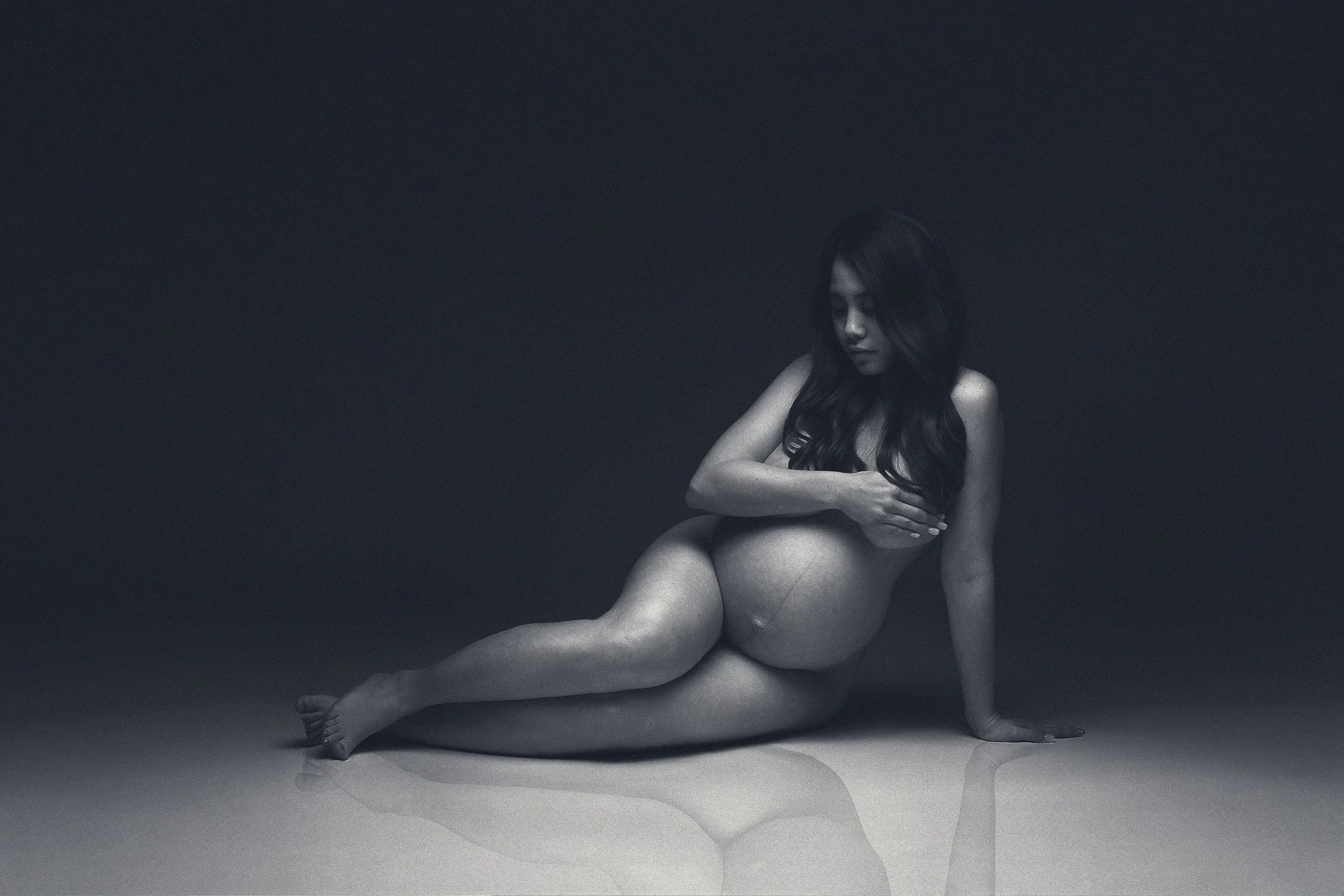 Black and white fine art maternity portrait of a pregnant woman seated on a studio floor, cradling her belly in dramatic low-key lighting. Photographed in Vancouver, WA.