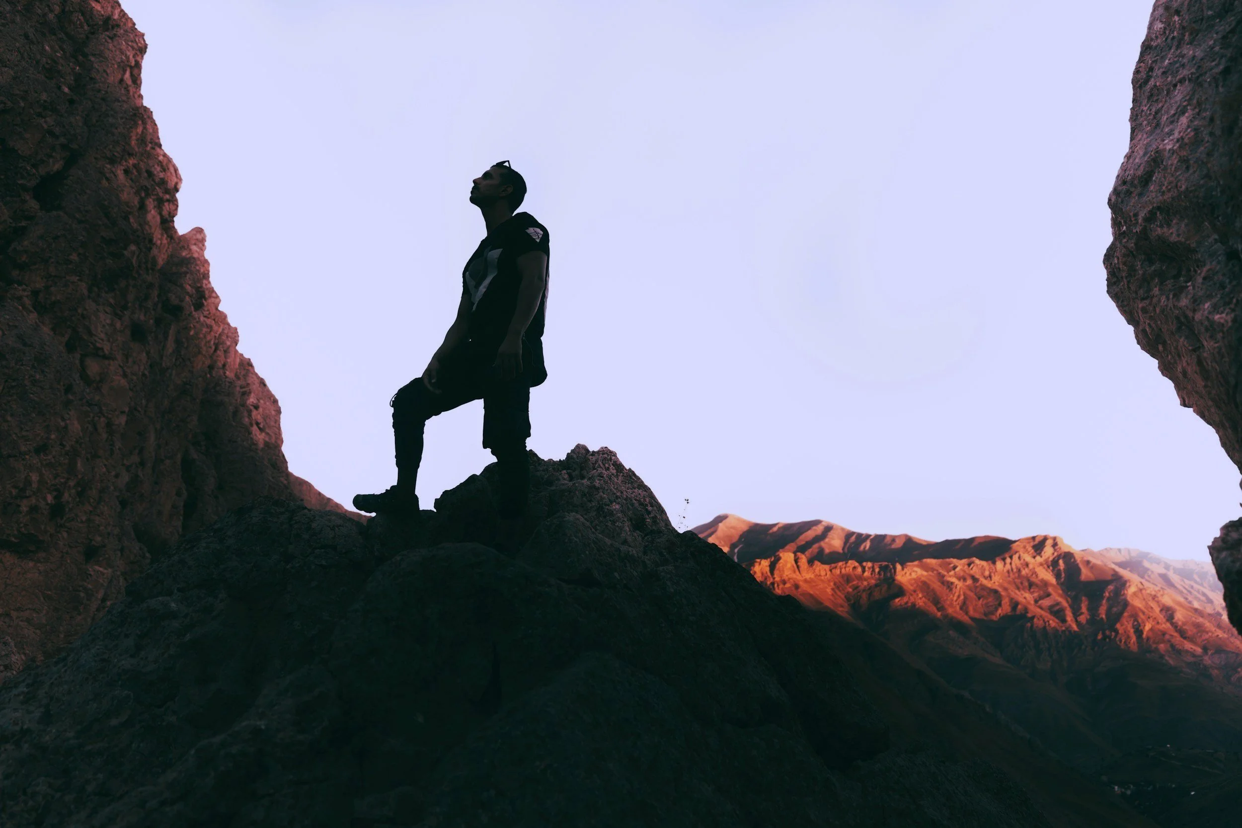 Silhouette eines Mannes, der auf einem Felsen in einer Bergregion steht, bei Sonnenuntergang oder Sonnenaufgang, mit umliegenden Bergen im Hintergrund.
