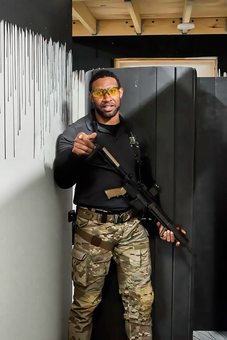 A man in camouflage cargo pants, black long sleeve shirt, holding a rifle, pointing at the camera, wearing yellow-tinted safety glasses, standing inside a building with a black wall, white drip paint design, and wooden ceiling.