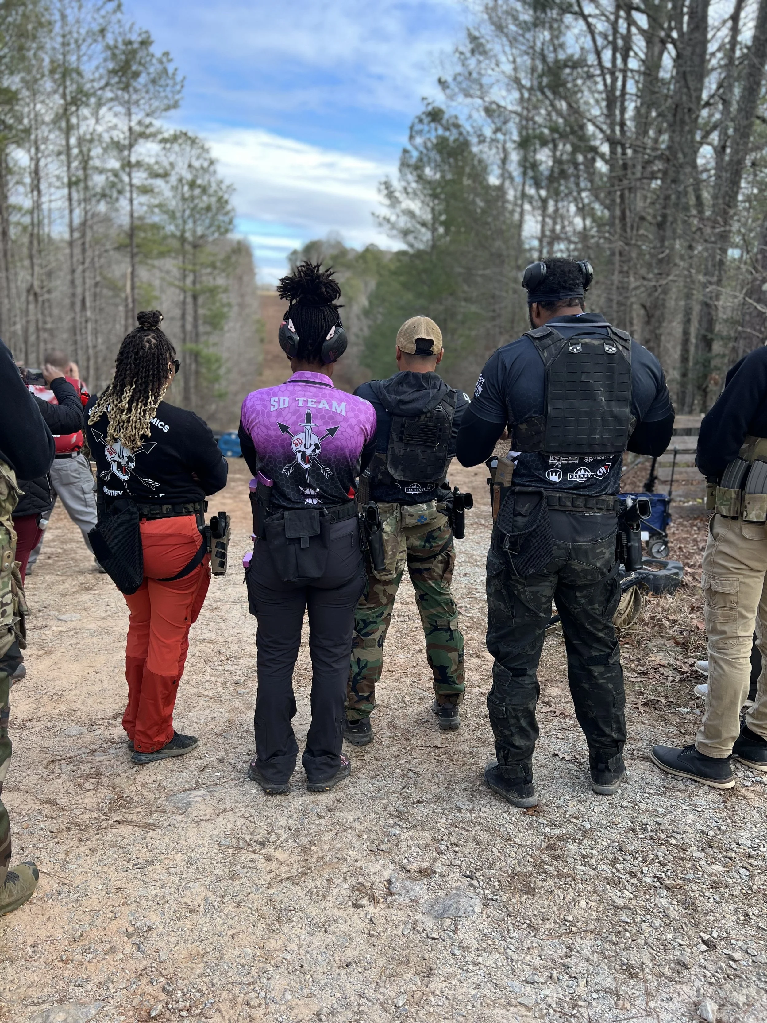 A group of people, including police officers and civilians, standing outdoors on a dirt path surrounded by trees, with some taking photos and others observing, under a partly cloudy sky.