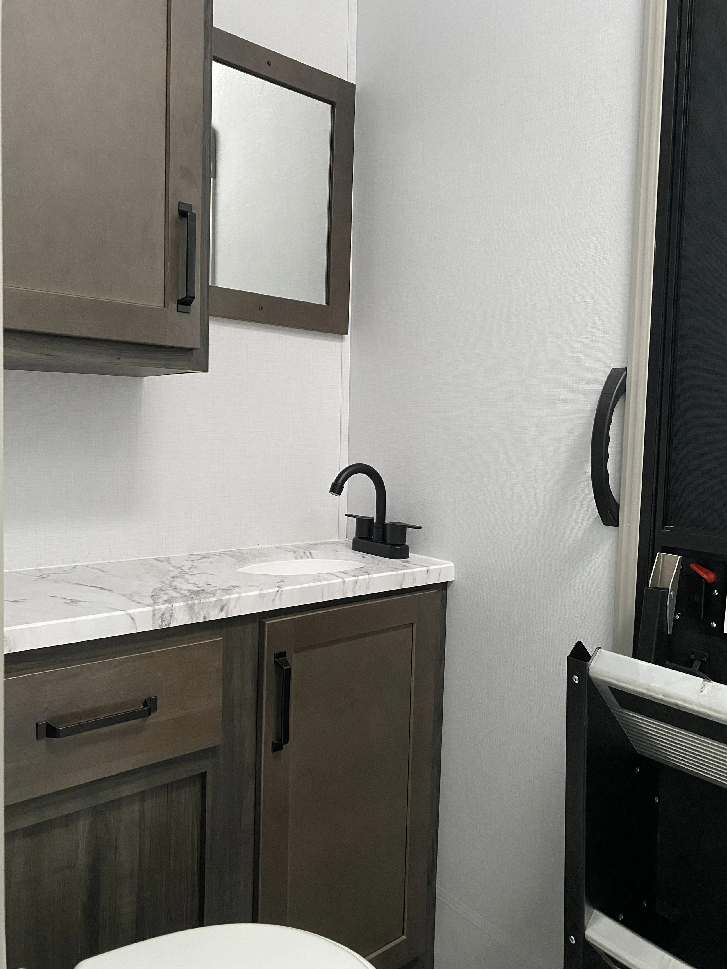 Small bathroom or utility room with a dark gray cabinet, a marble countertop with an almond-shaped sink, a black faucet, wall-mounted mirror, and a white wall. Part of a black shelving unit or cabinet is visible on the right.