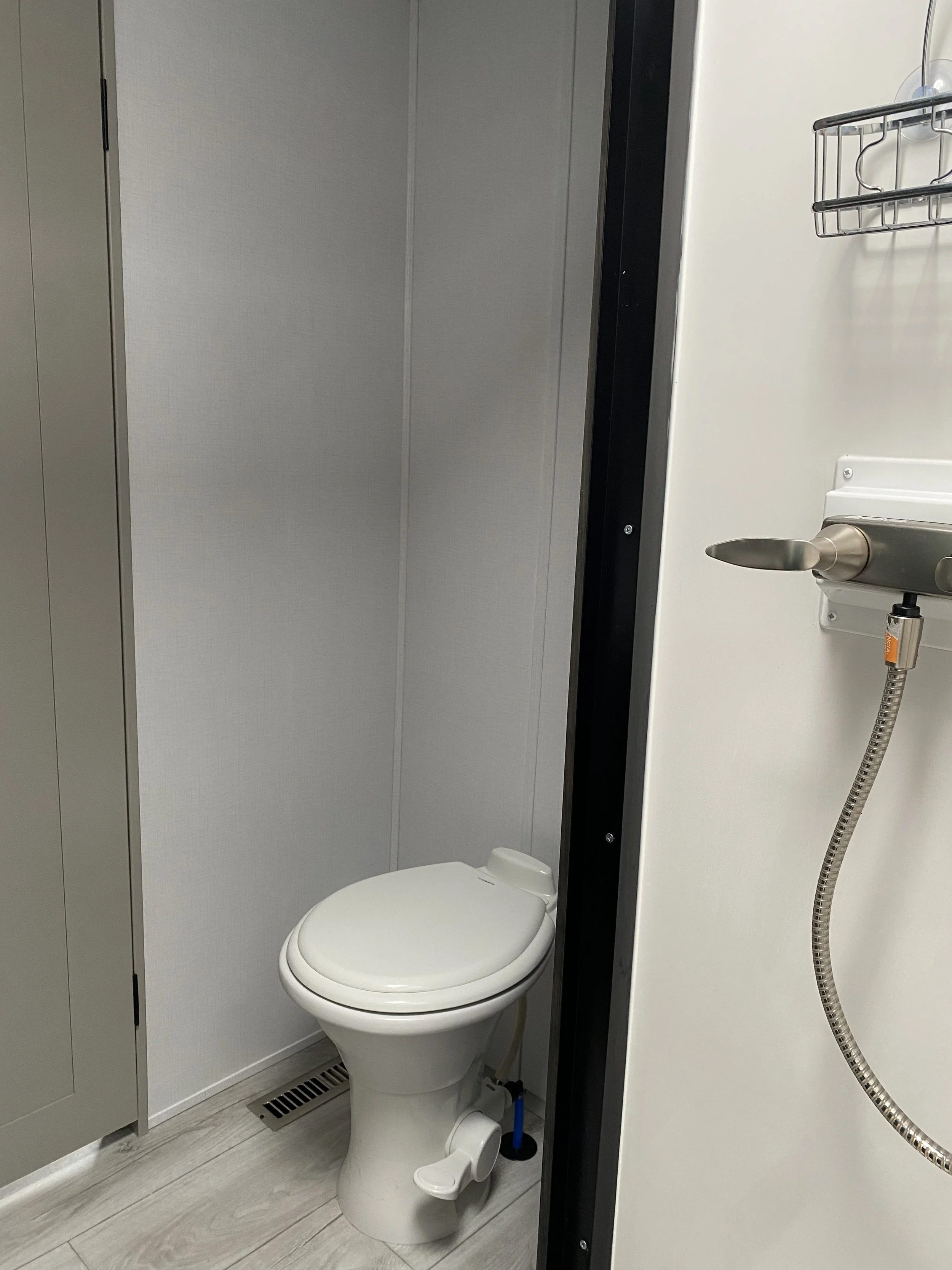 A small bathroom with a toilet and a shower area. The toilet is white, and there is a vent on the floor. The shower area has a handheld shower head attached to the wall and a wire shelf.