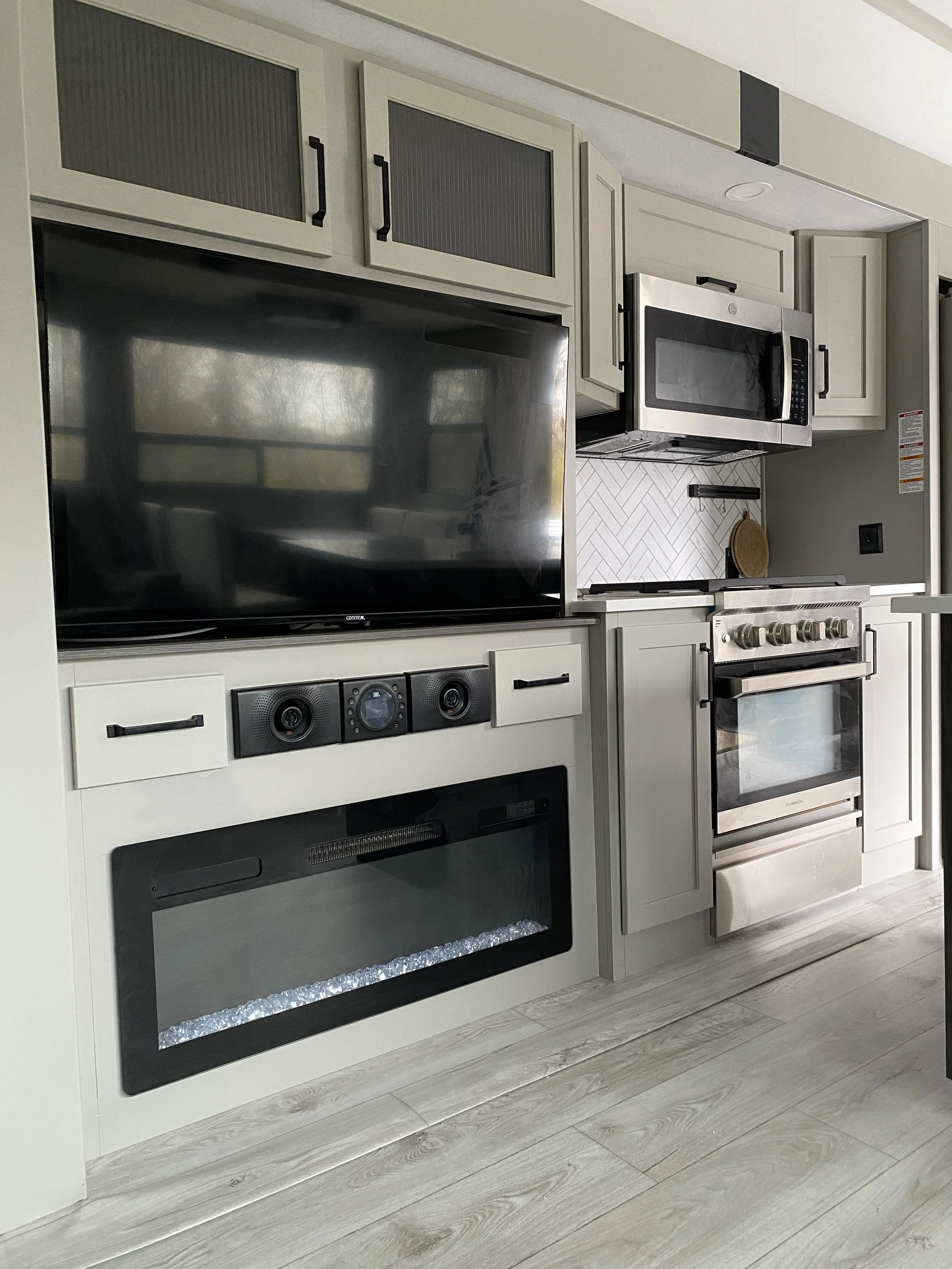 Modern kitchen with white cabinetry, stainless steel microwave, oven, large flat-screen TV, and electric fireplace, with light wood flooring.
