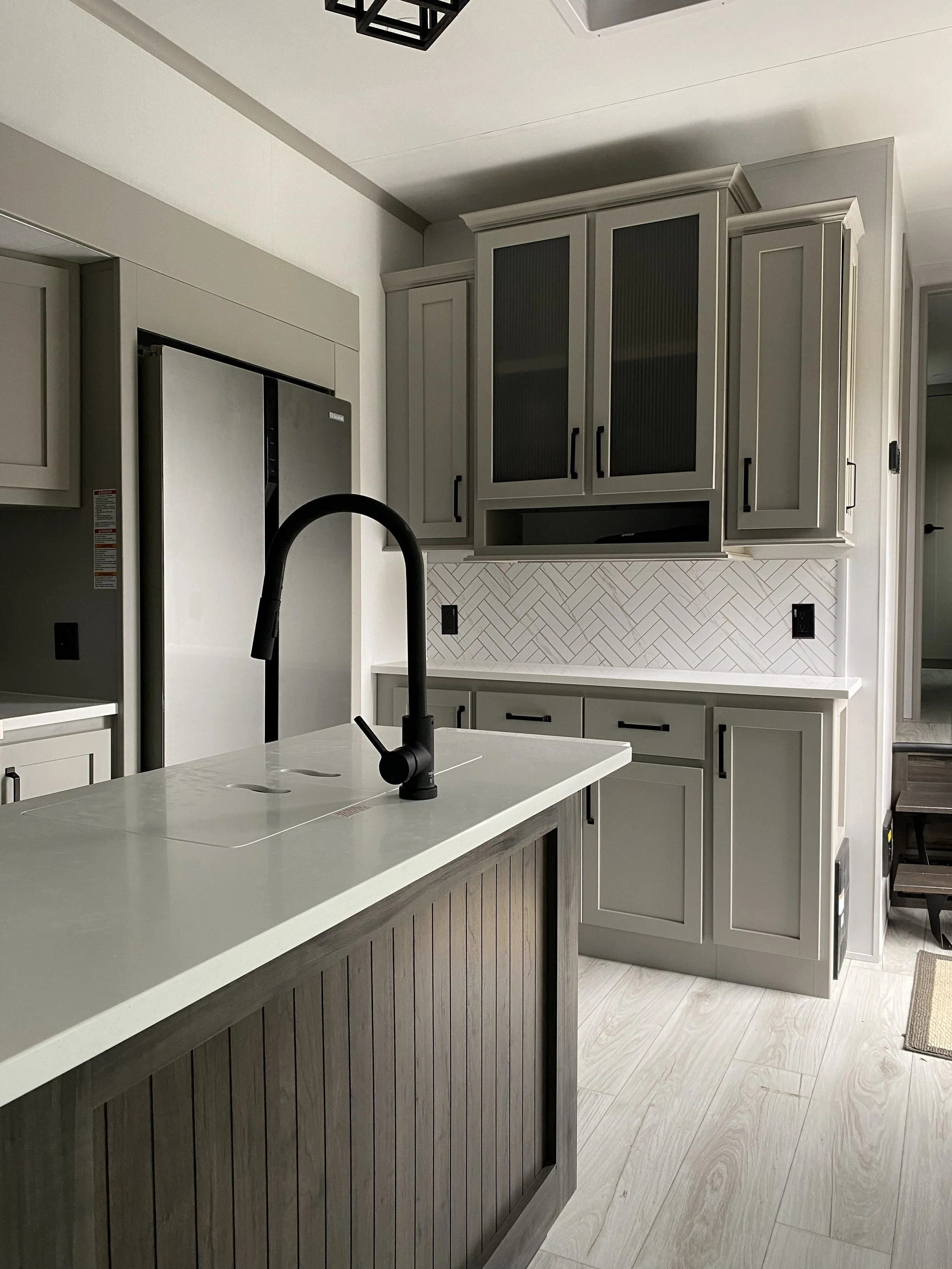 Modern kitchen with light gray cabinets, a white countertop, black fixtures, and a patterned tile backsplash. There is a dark wood island in the foreground and stainless steel appliances.