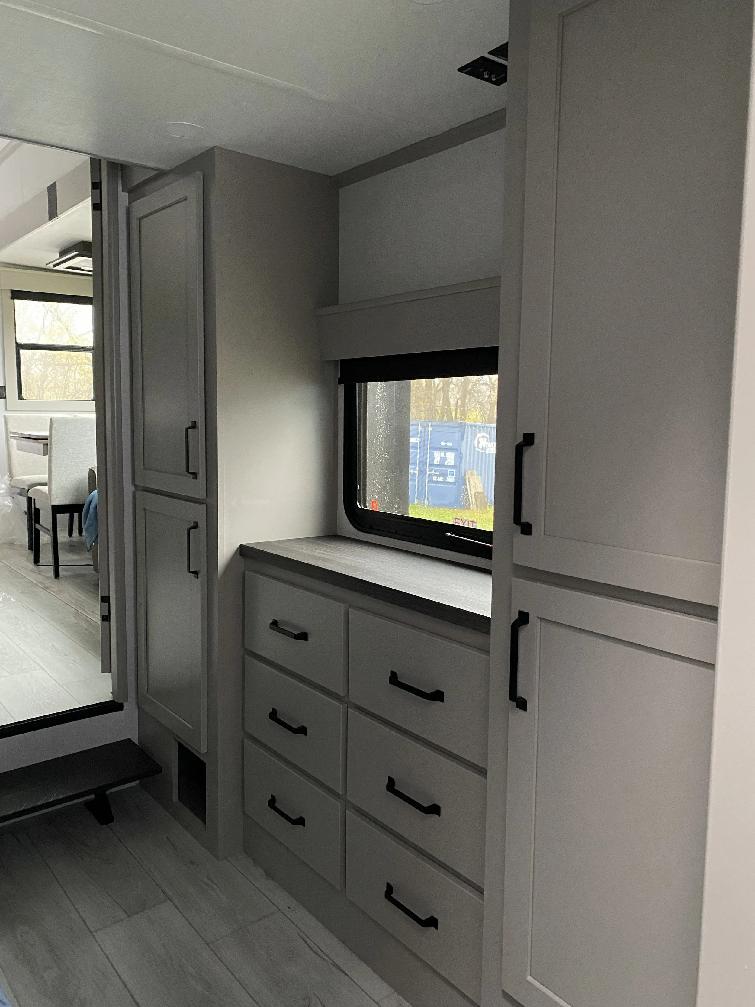 Interior view of a modern RV or tiny home with built-in cabinetry, a window with a black roller shade, and a glimpse of a dining area outside.