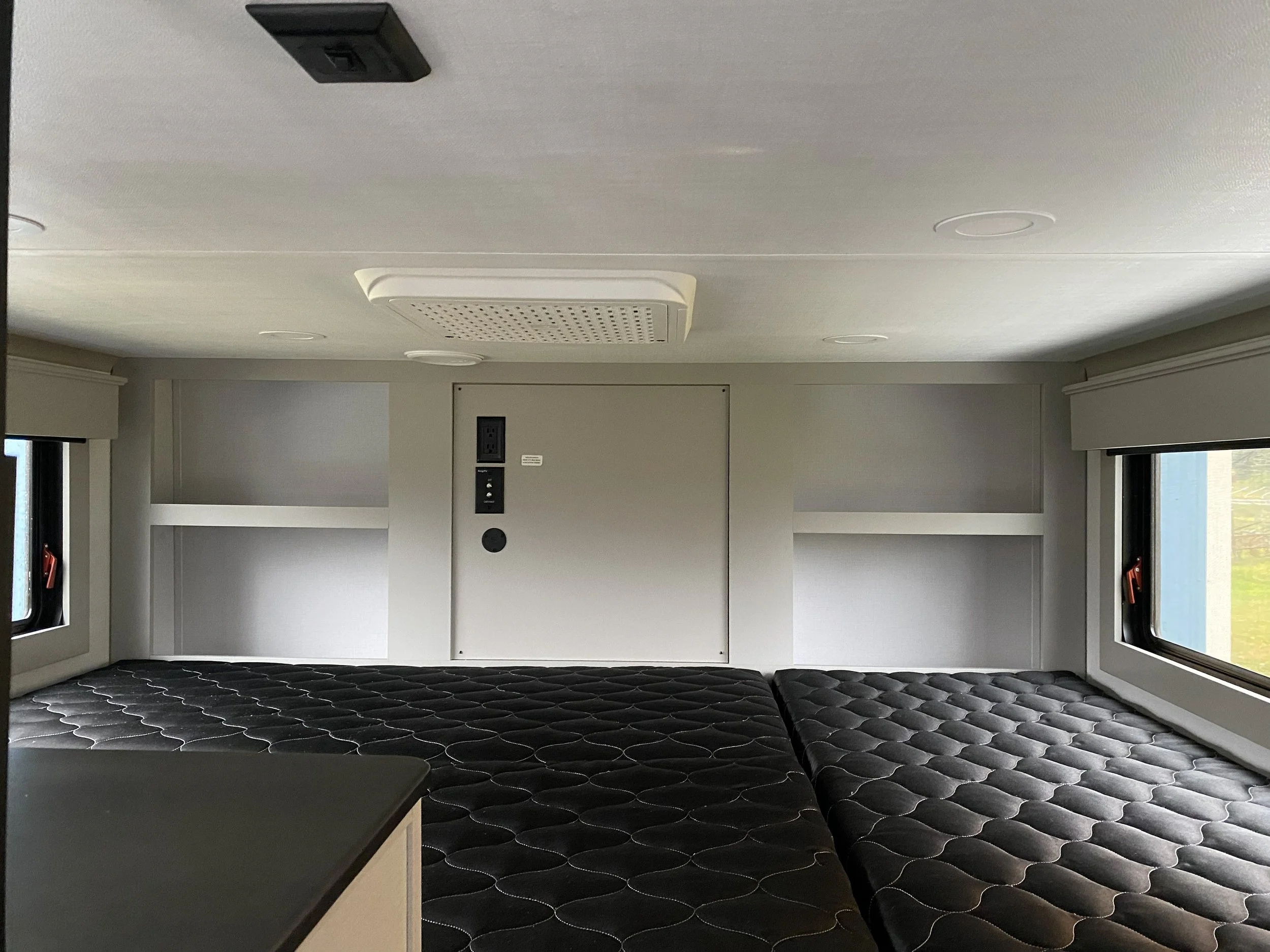 Interior of a room with two twin beds, one on each side, with black mattresses and white piping, and two windows on either side showing outside scenery. The ceiling contains a light fixture, an air vent, and a black speaker.