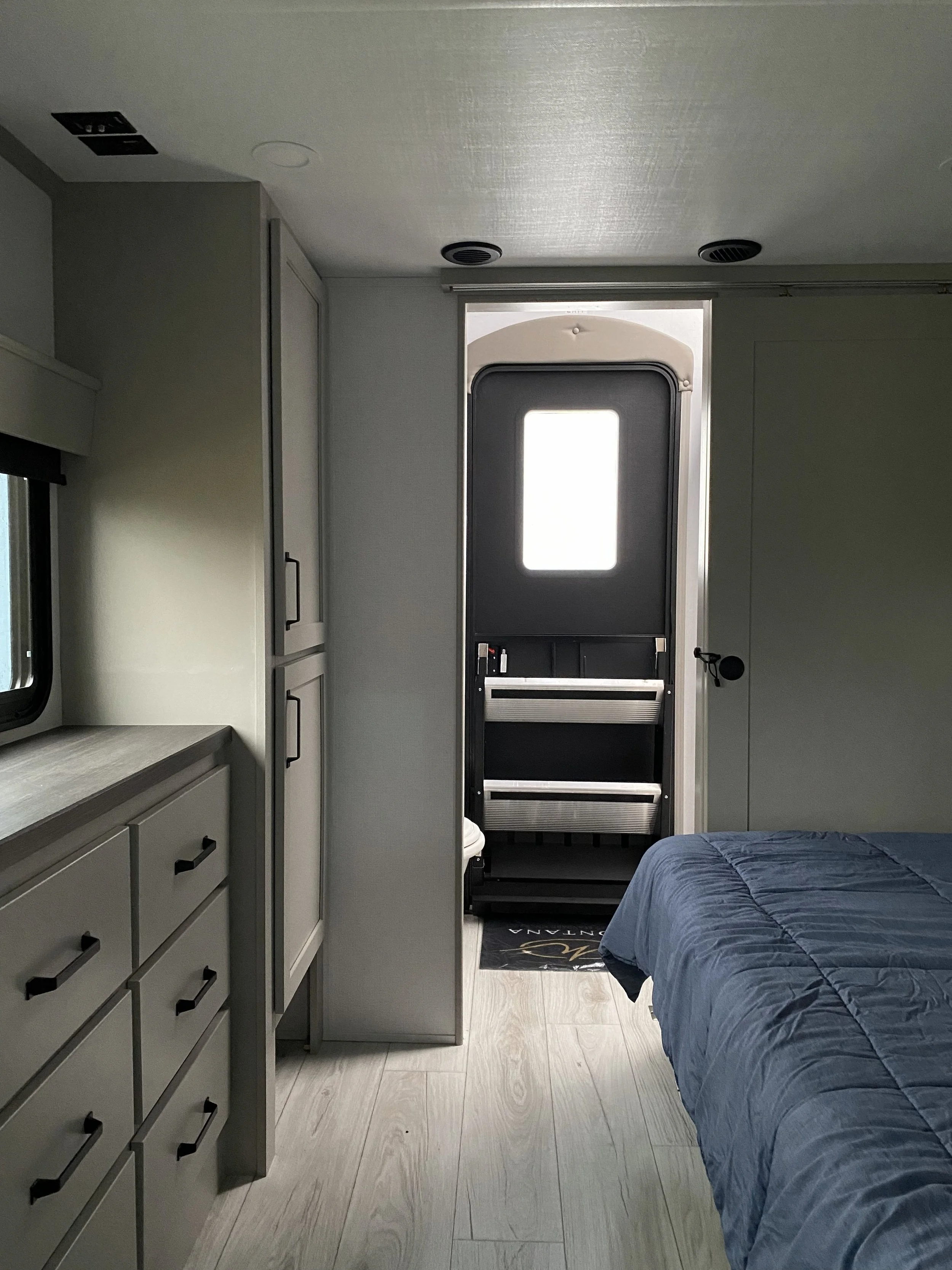 Interior of an RV bedroom showing a bed with dark blue bedspread, a four-drawer dresser on the left, and a door leading outside with a black frame and a small window in the center. Small ceiling vents and a window with a blind are also visible.