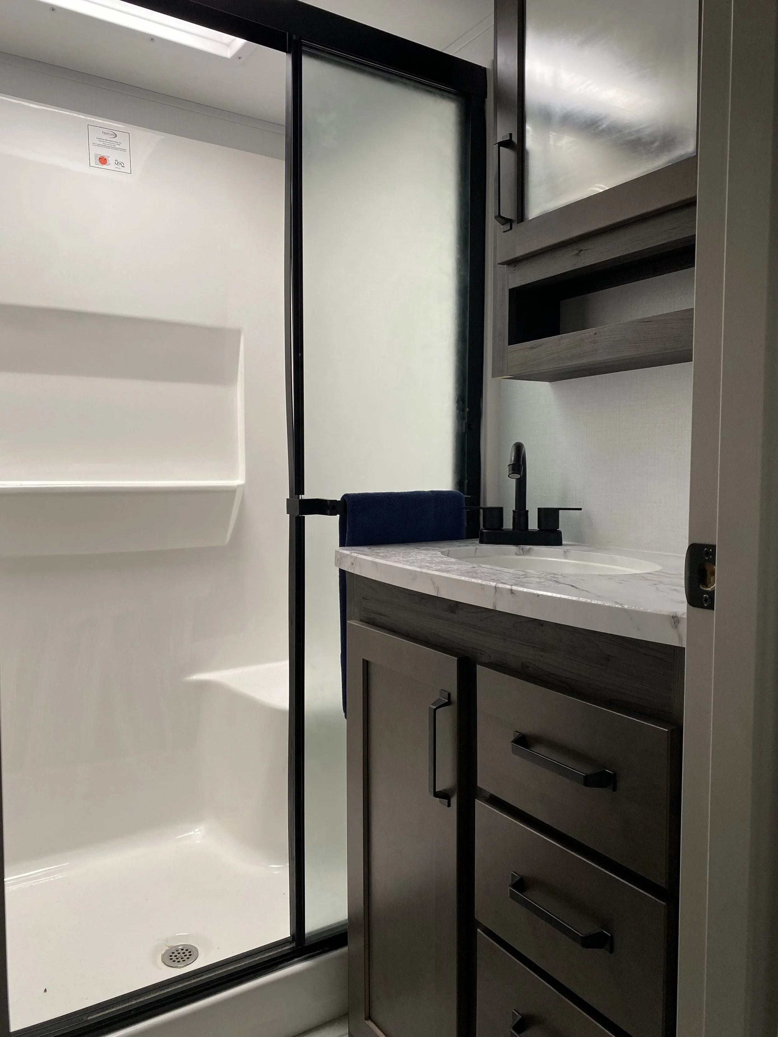 Small bathroom with a shower stall on the left, a white marble countertop with black faucet in the center, and dark wood cabinets below and above the sink on the right.