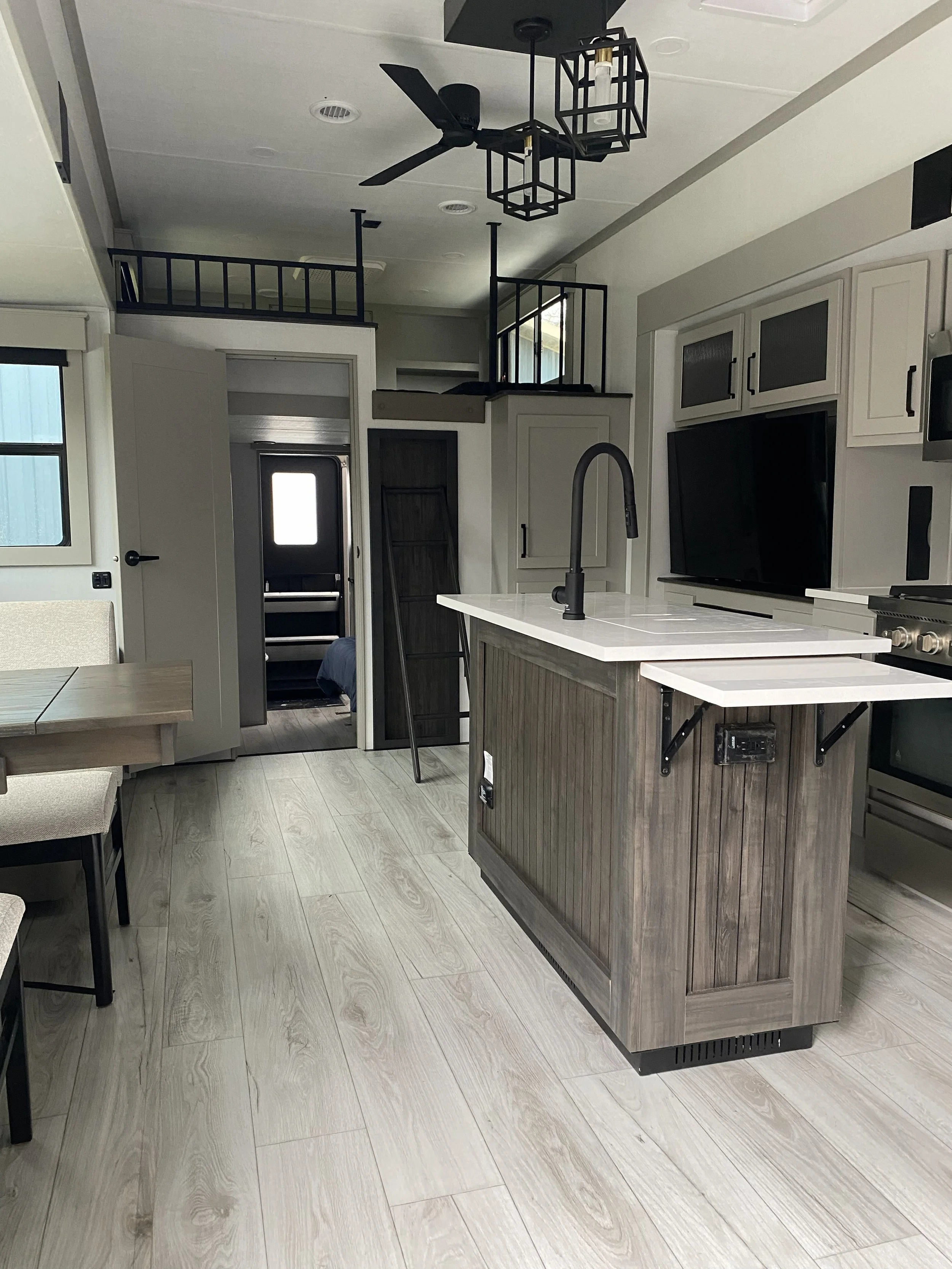Modern kitchen with light wood flooring, central island with a white countertop, black faucet, and gray cabinetry. Open door to a small entryway and stairs in the background. Ceiling fan and modern light fixtures.