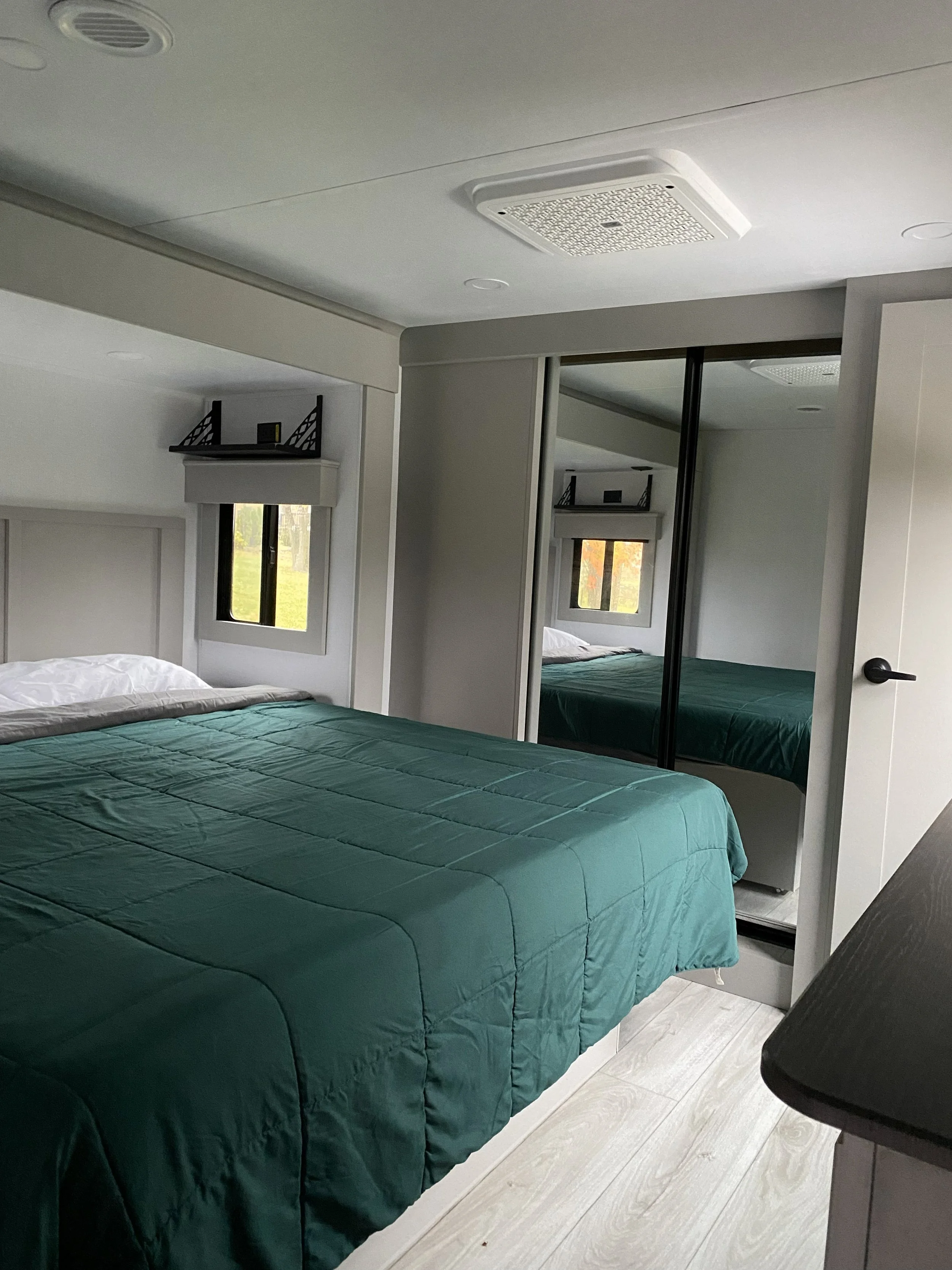 A bedroom with a bed covered in a green quilt, small window, mirrored closet doors, and ceiling vent.