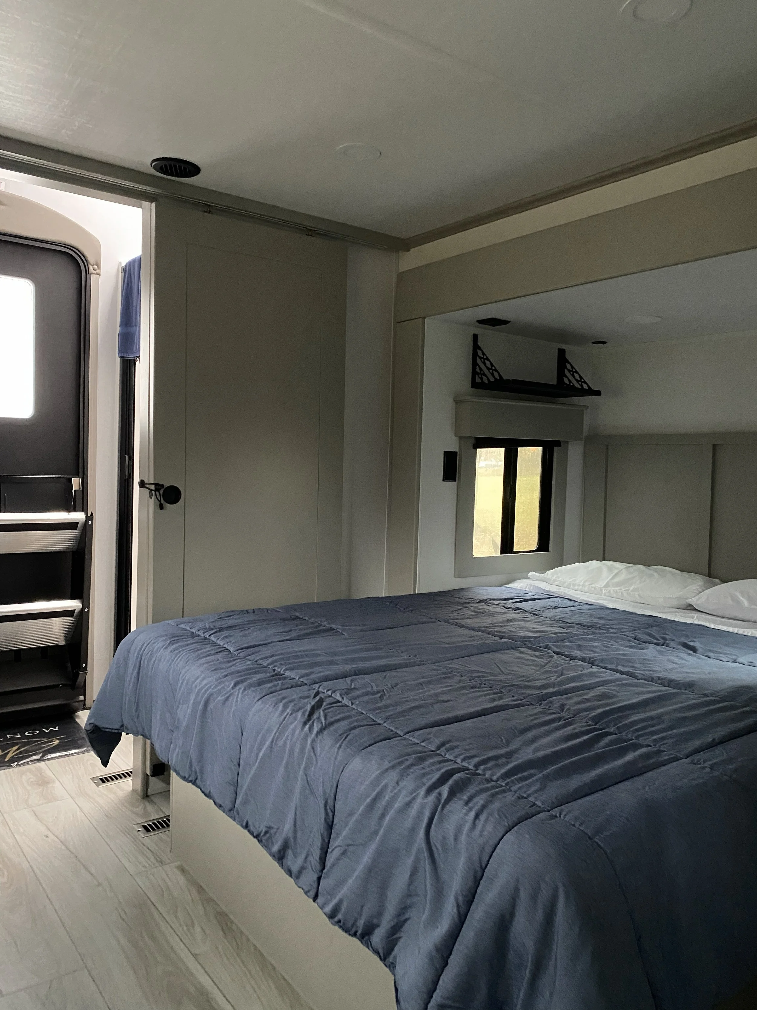 Interior of a bedroom in a camper with a bed, window, and shelves.