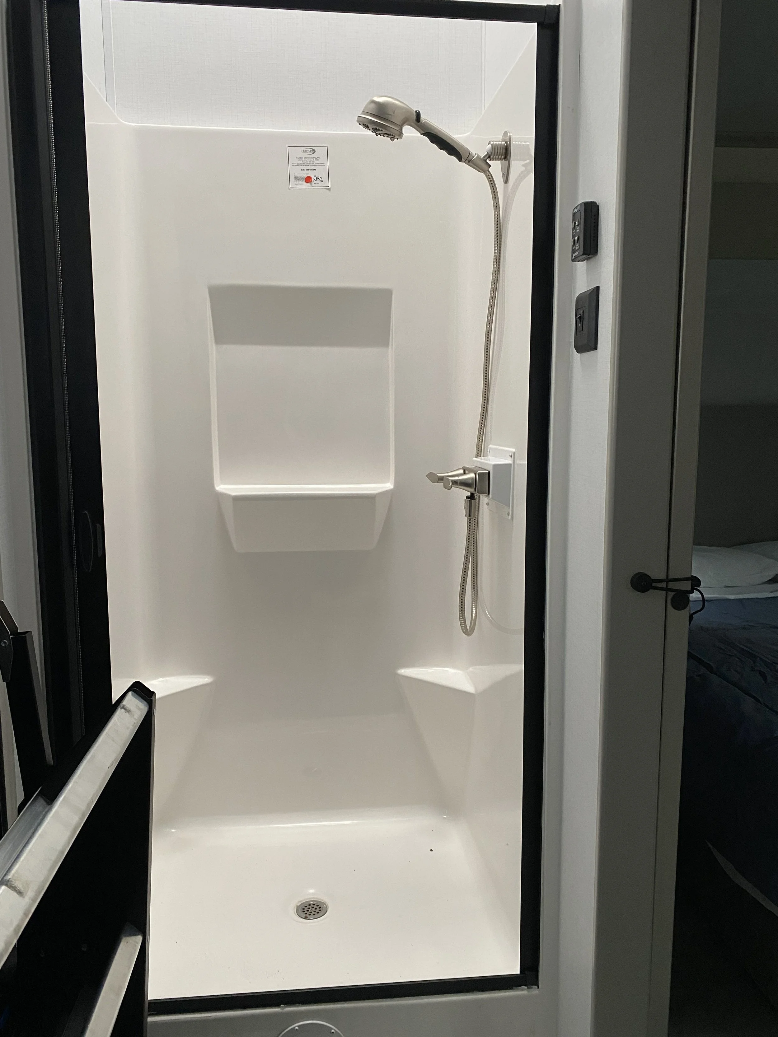 A white shower with a handheld showerhead, built-in shelving, and a drain in the floor. The shower is enclosed with black framing on the edges.