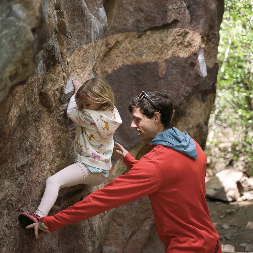 A young girl wearing a colorful dinosaur-printed hoodie and beige leggings is climbing a rock wall outdoors. A man in a red jacket and sunglasses on his head is assisting her by showing her foot placements and providing support.