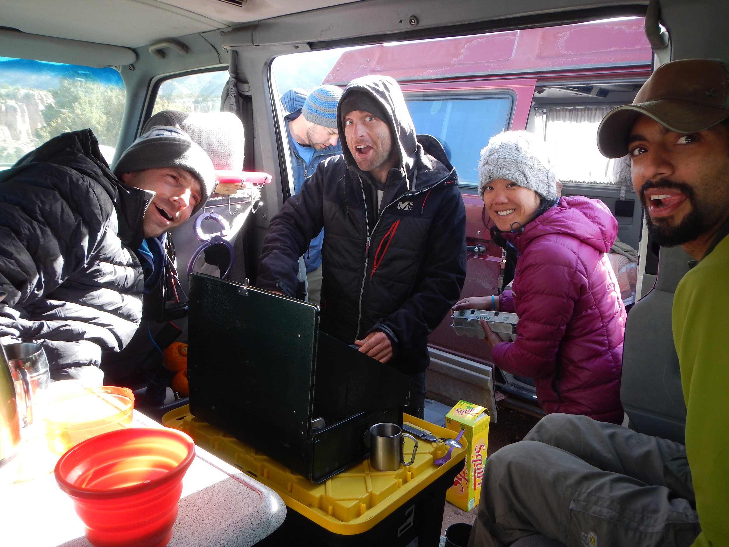 Group of five people inside a camper van, wearing outdoor jackets and beanies, with a kitchen setup and a view of a red van outside.