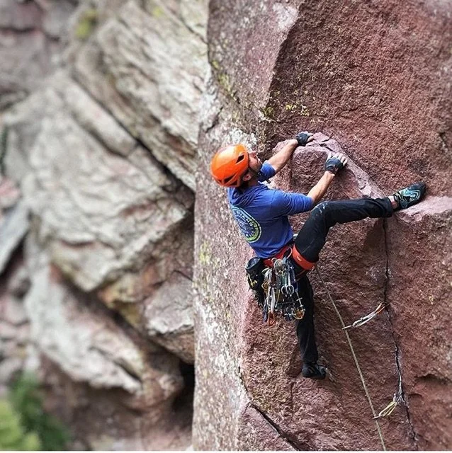 A person rock climbing on a vertical red and brown cliff face, wearing an orange helmet, blue shirt, harness, and climbing gear.
